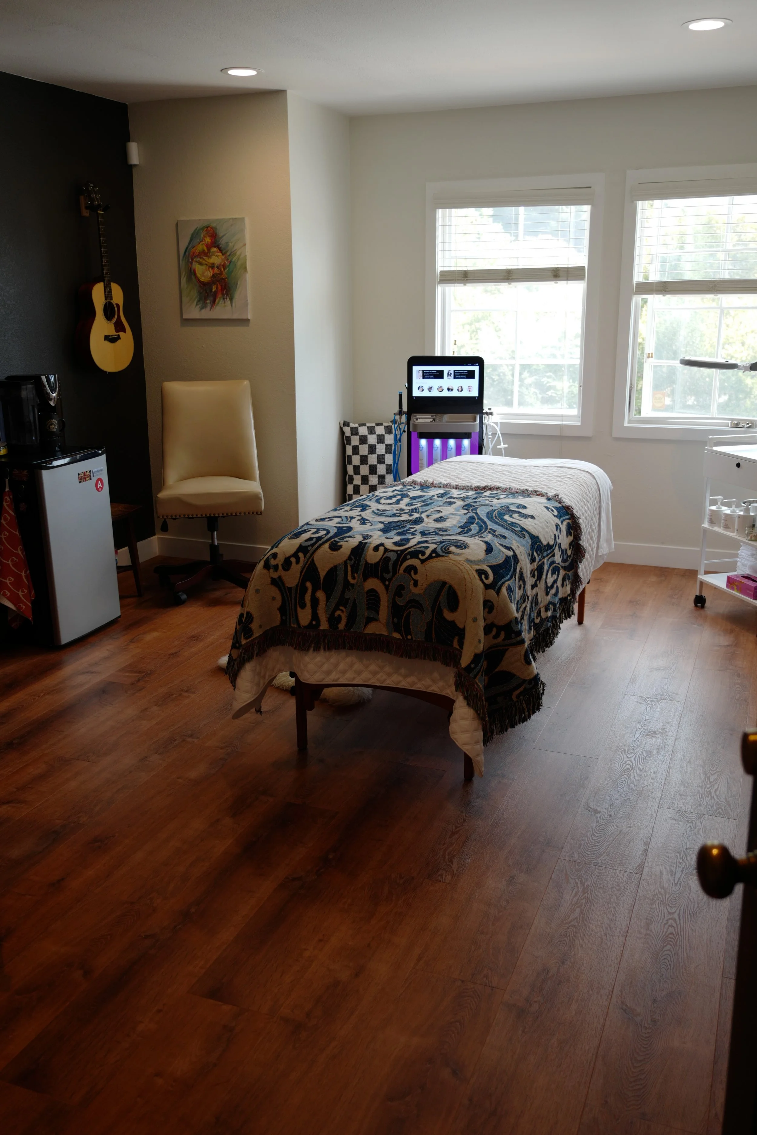A room with wooden flooring, a massage table with a patterned blanket, a beige chair, a guitar hanging on the wall, a colorful abstract painting, and a window letting in natural light.