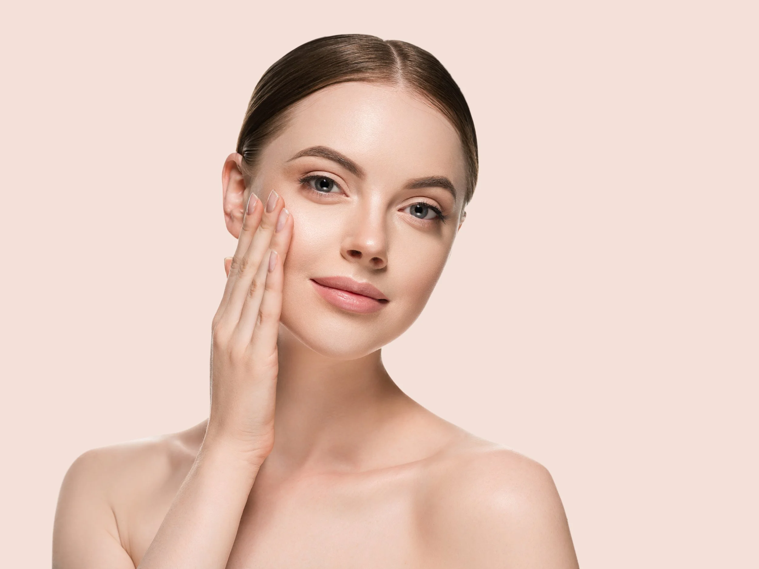 Close-up of a woman with clear skin and brown hair, gently touching her cheek with her fingers, against a light pink background.