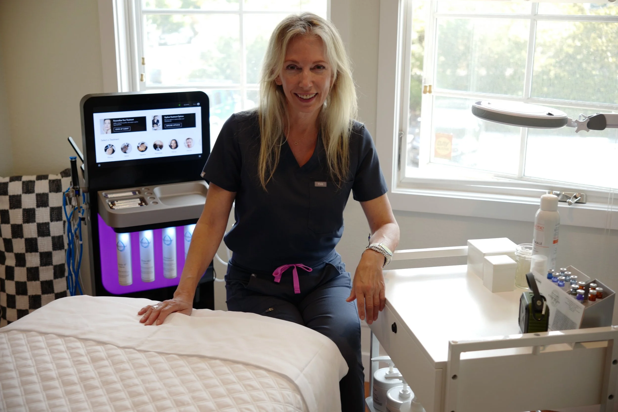 A smiling female healthcare professional sitting on the edge of a hospital bed in a medical office, with medical equipment and supplies nearby and a large window in the background.