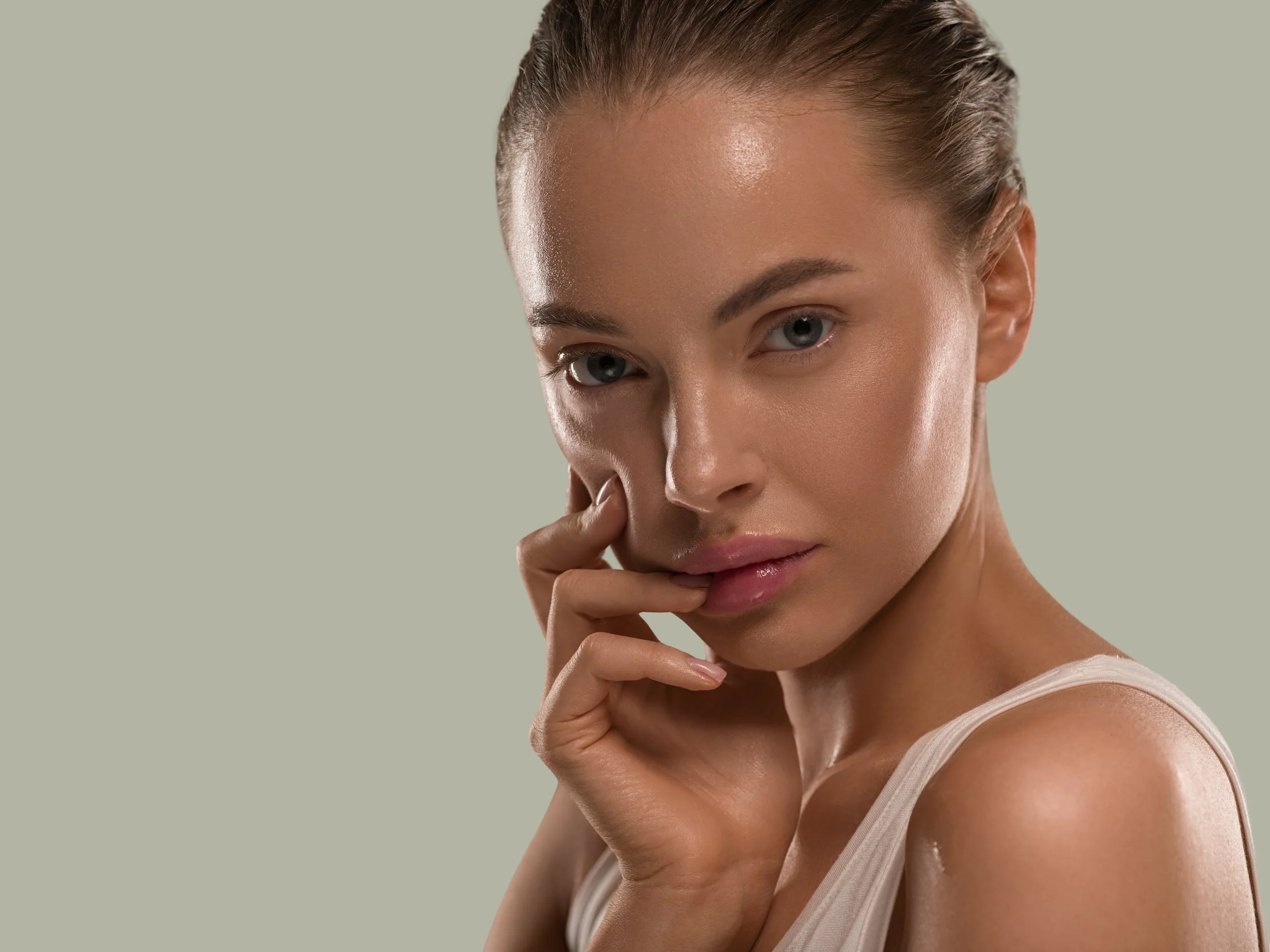 A woman with her hand near her face, looking directly at the camera, with smooth skin and slicked-back hair.