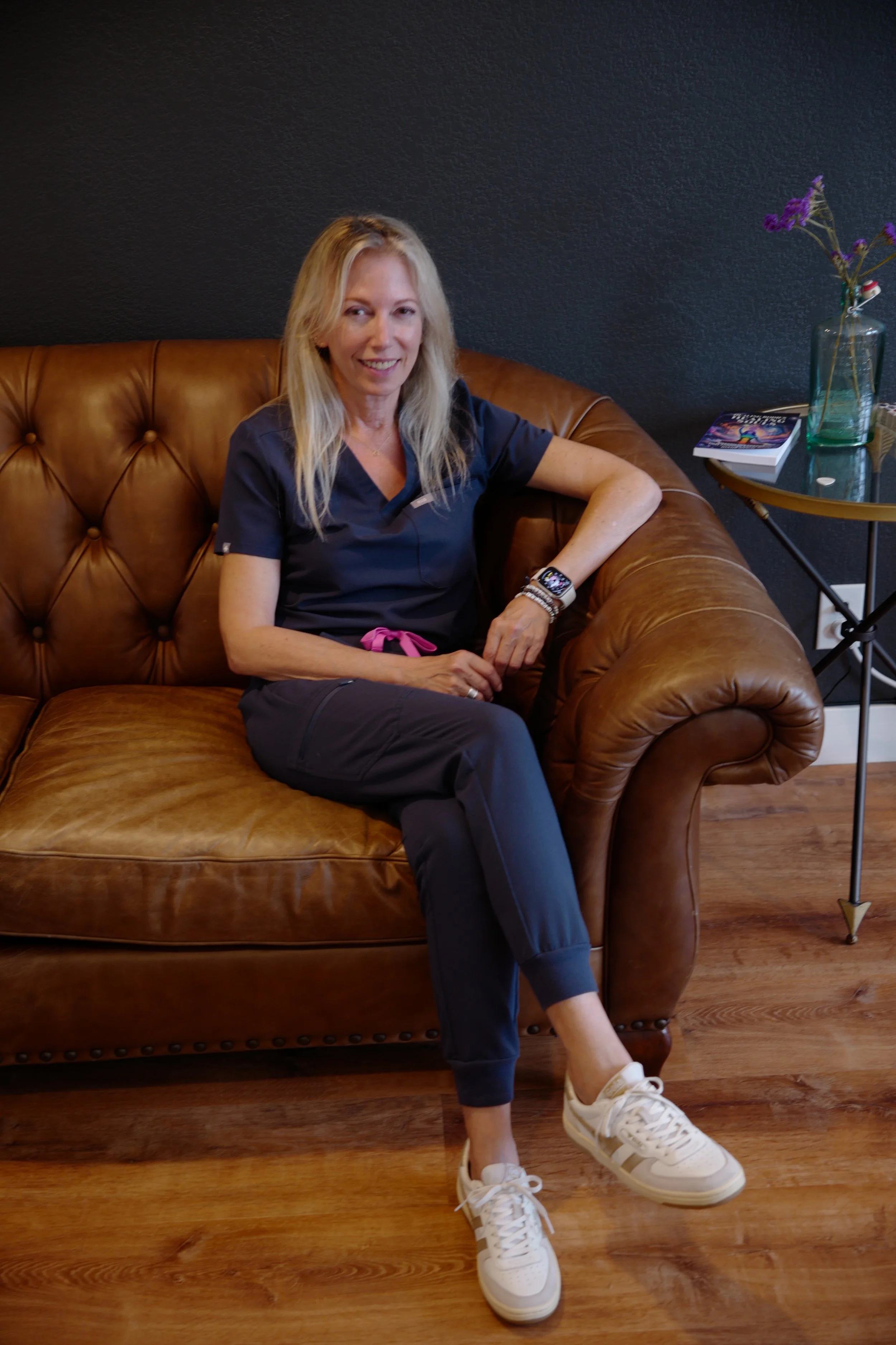 A woman with blonde hair wearing a navy scrubs sitting on a brown leather couch, smiling, with a dark wall behind her and a side table with a book, a magazine, a glass bottle with flowers, and a few small objects nearby.
