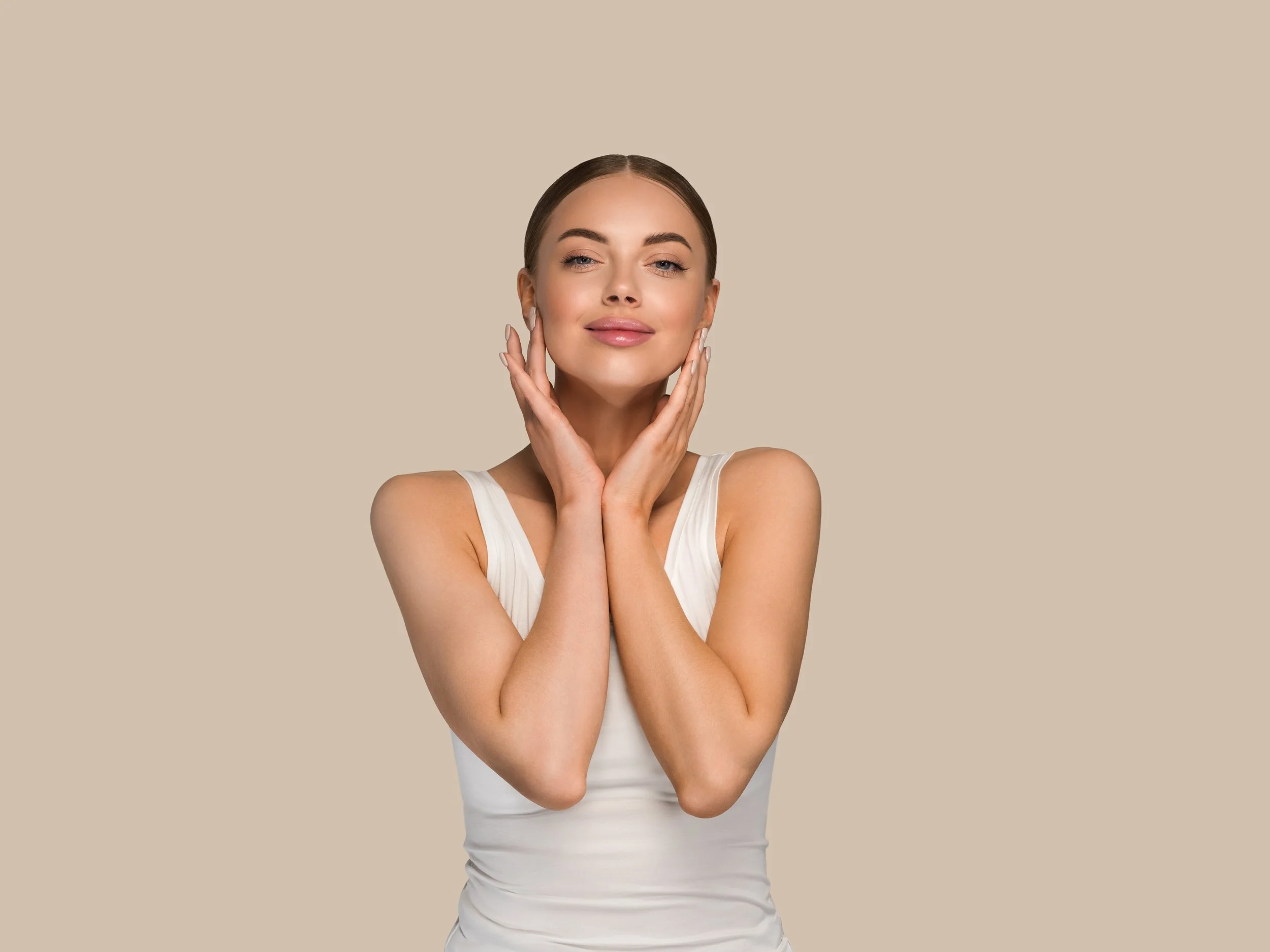 A woman with dark brown hair and fair skin touching her face gently with both hands, wearing a white sleeveless top, and smiling softly at the camera against a plain, beige background.