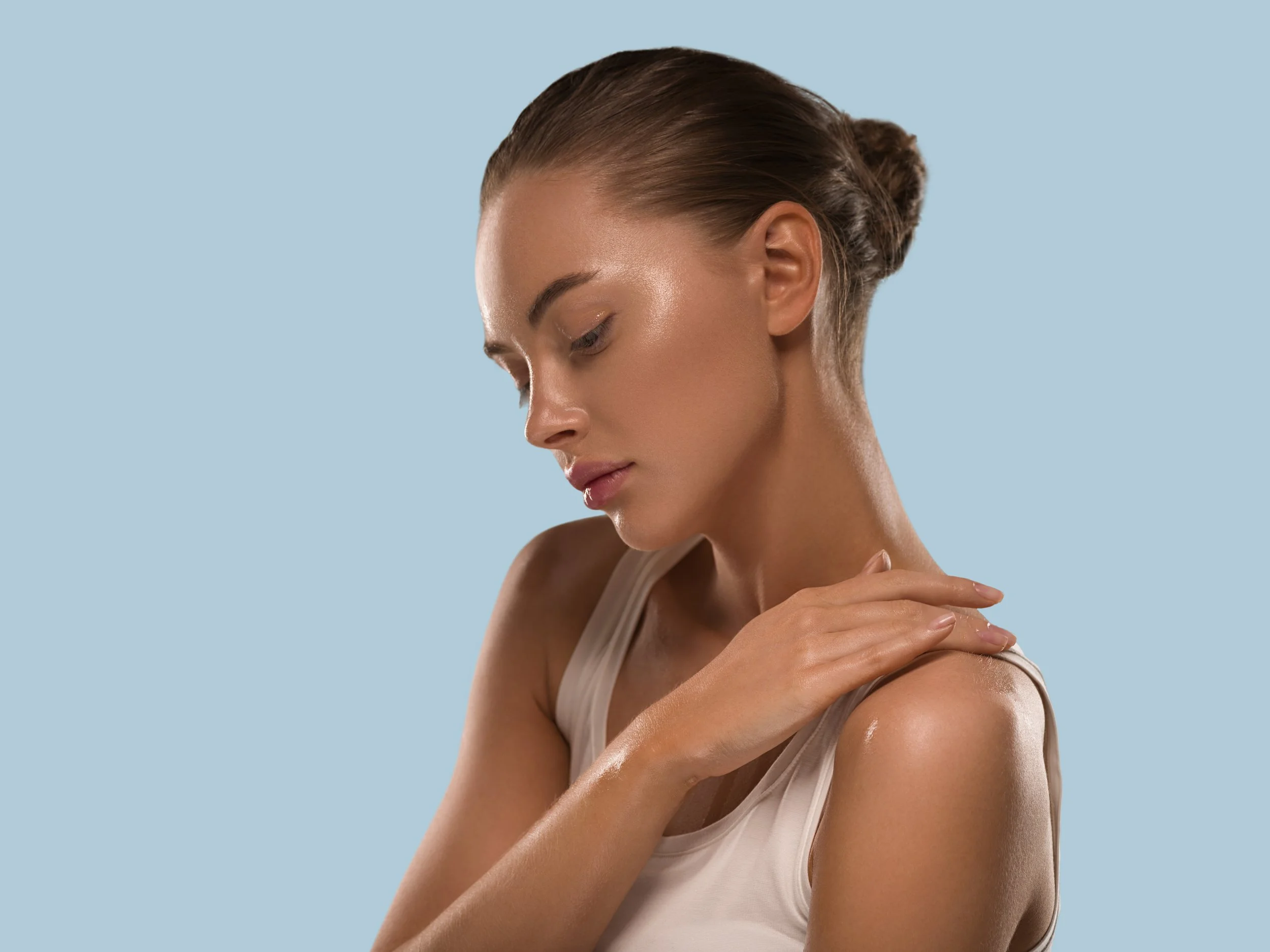 A woman with her eyes closed, her hair in a bun, wearing a beige tank top, and touching her shoulder against a light blue background.