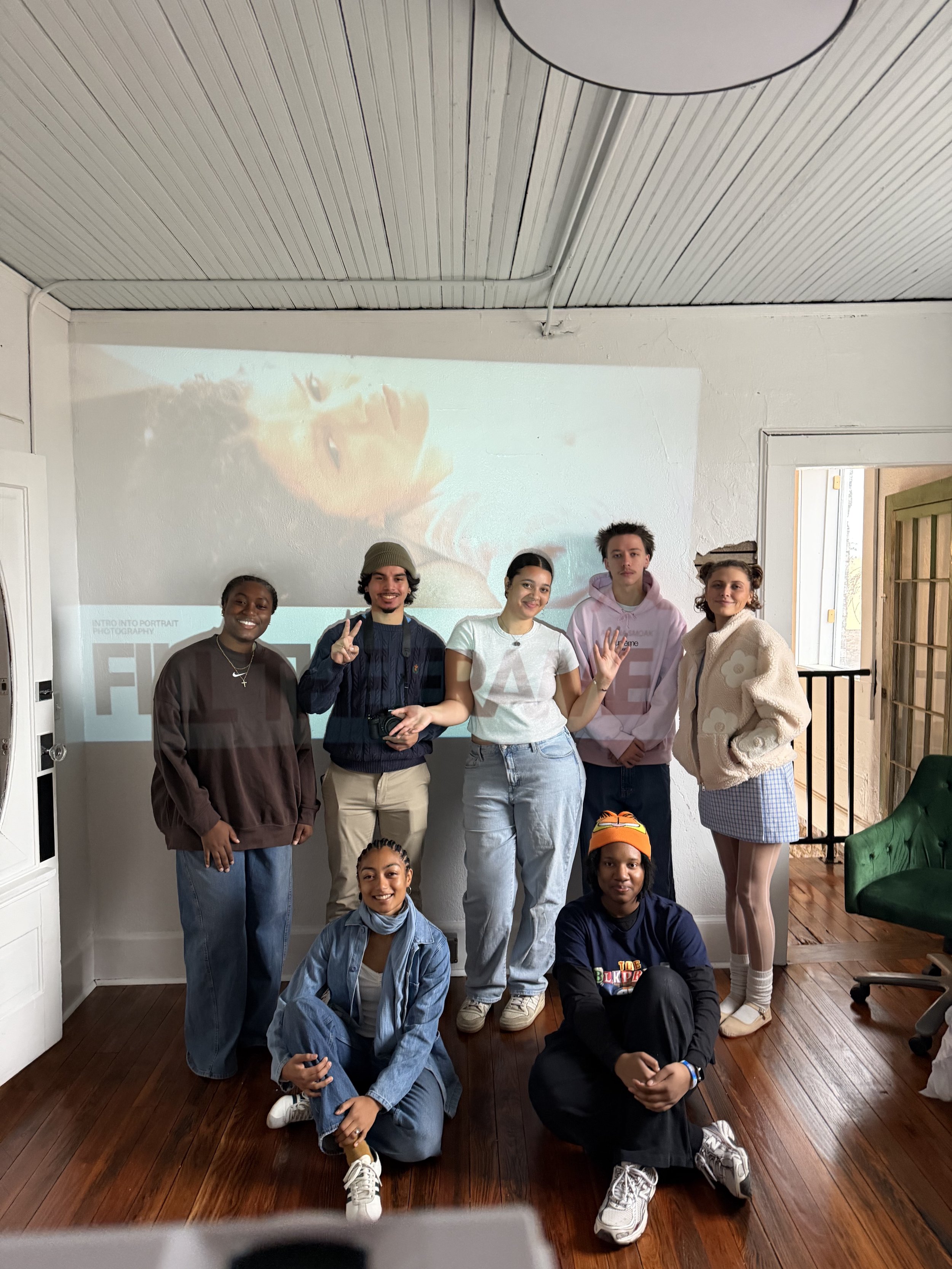Group of ten young people posing indoors in front of a wall with a projector screen showing a portrait of a woman. They are standing and sitting on a wooden-floored room, dressed casually, smiling, and making various gestures.