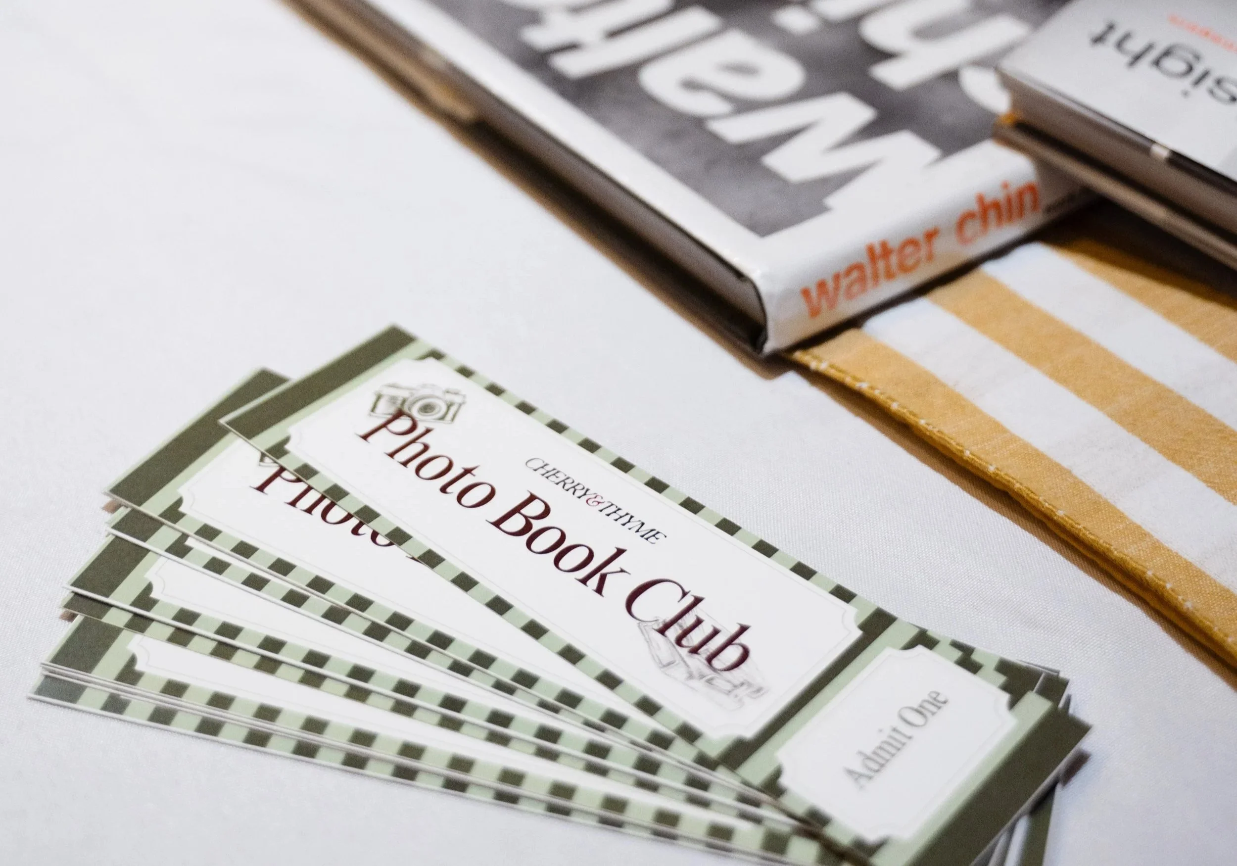 A stack of green and white checkered coupons with the words "Photo Book Coupon" and a small image of a camera on them, placed on a white surface. Behind the coupons, there are several books or magazines, with the top one having a black and white cover and orange text reading "Walter Ching".