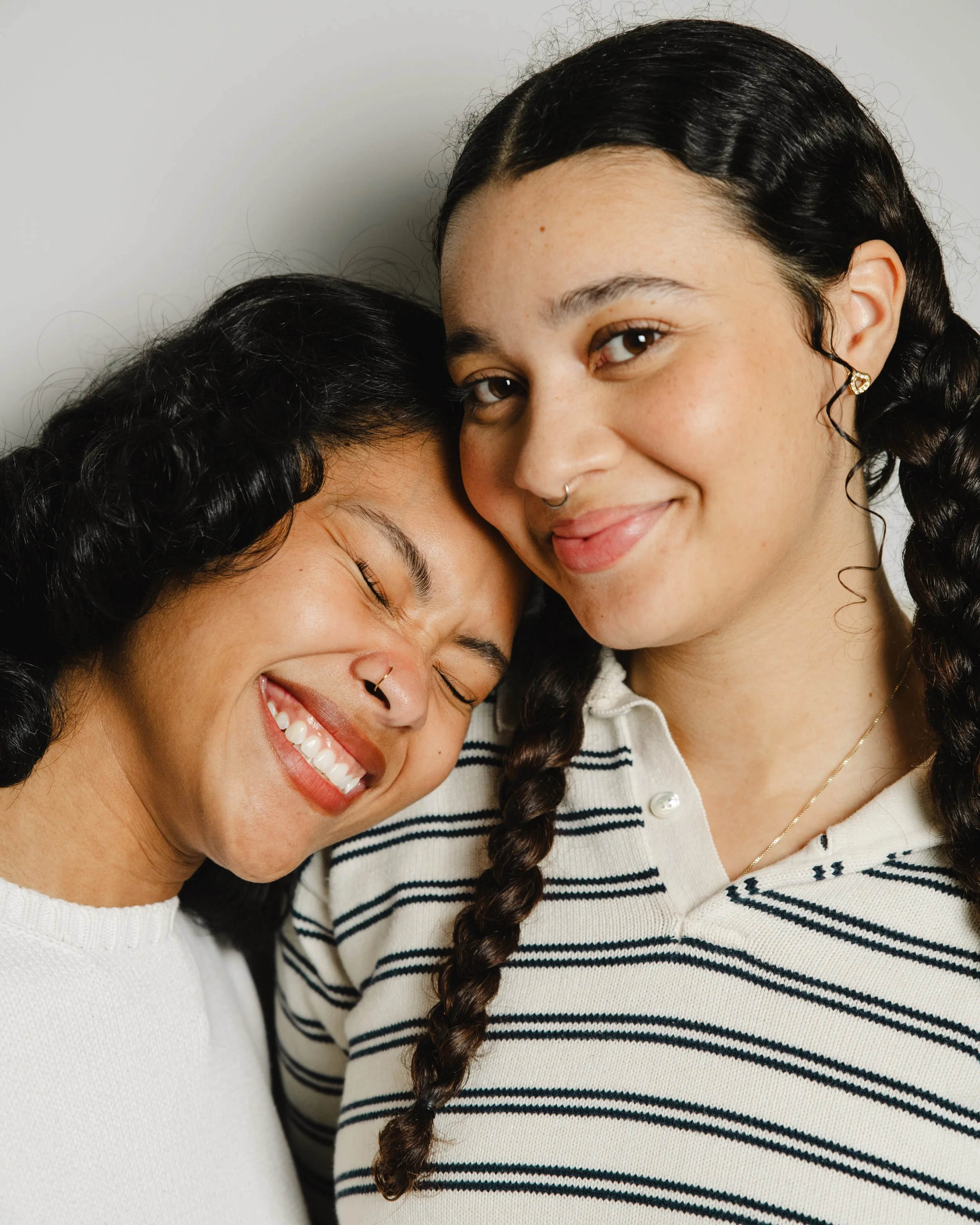 Two women with dark hair and freckles smiling and hugging, one woman wearing a striped shirt and gold earrings, the other in a white sweater.