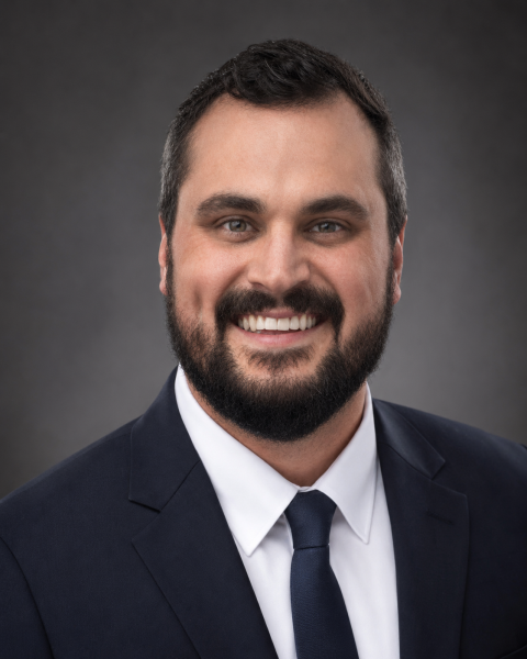 A smiling man with dark hair and a beard, wearing a navy blue suit, white shirt, and dark tie, against a gray background.