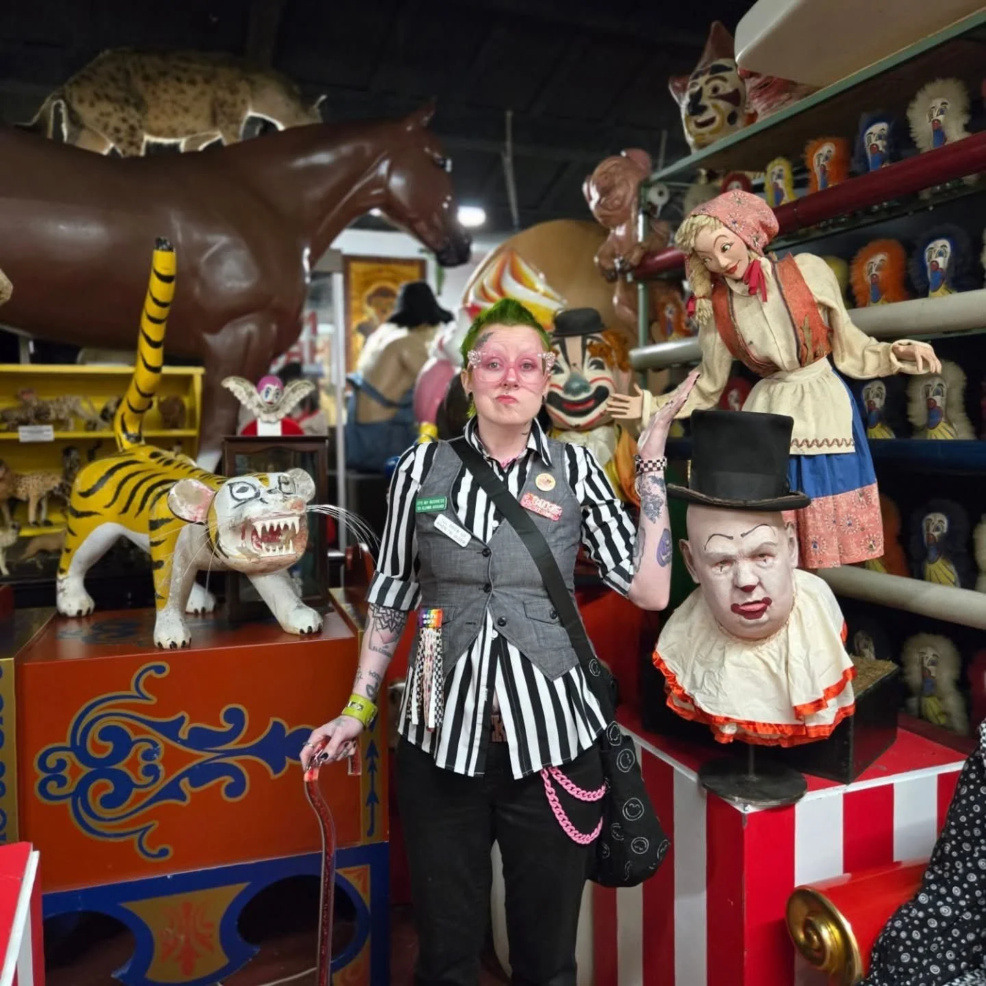 Person with pink face makeup, green hair, and tattoos standing among vintage clown and animal figurines in a circus memorabilia store.