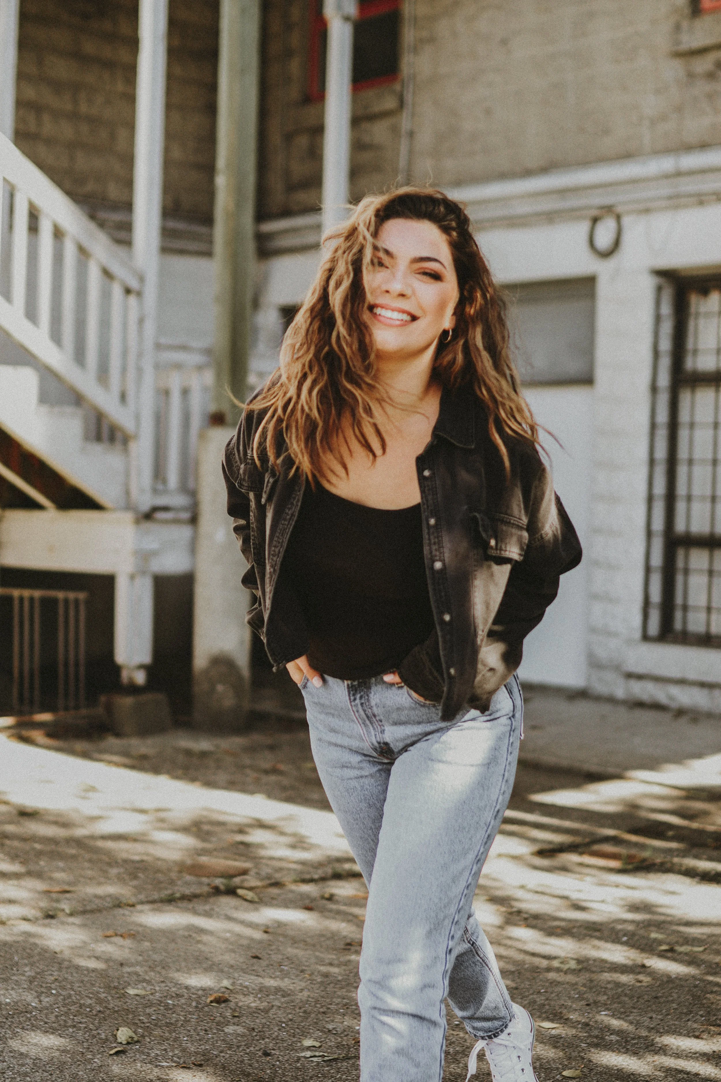A young woman with wavy hair smiling, walking outdoors in front of a building with stairs, wearing a black top, a black denim jacket, and light blue jeans.