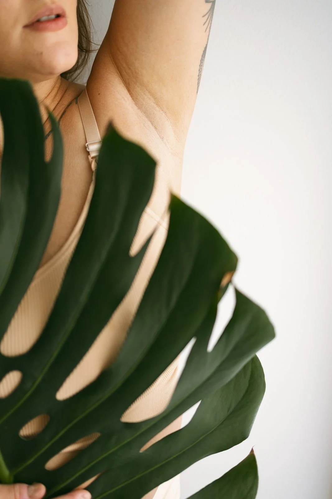A woman with a tattoo under her raised arm, dressed in beige, partially obscured by a large green Monstera leaf, with a neutral background.