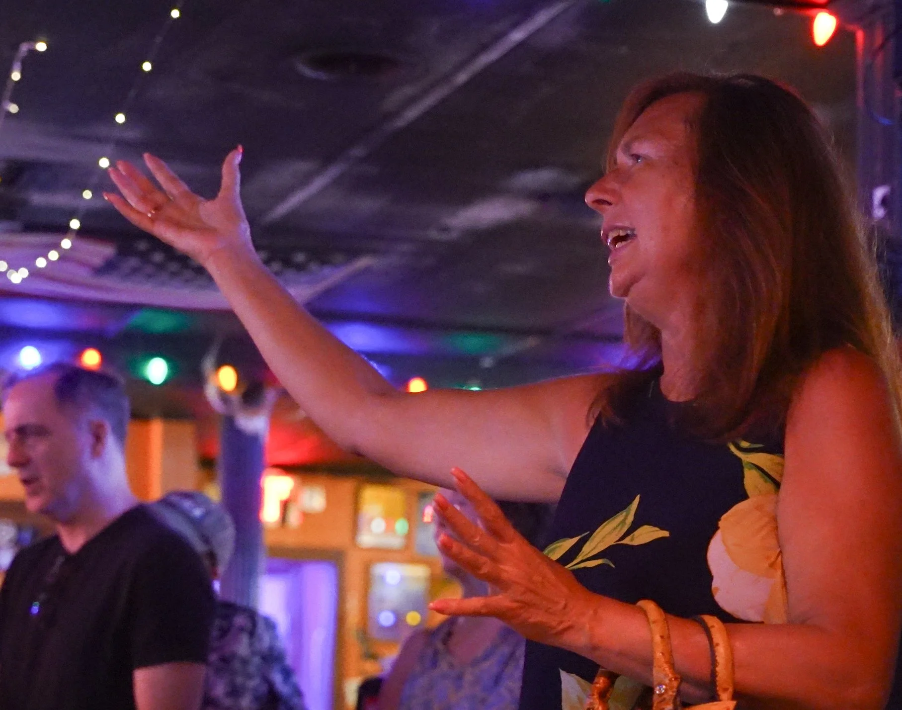A woman with long hair and a floral dress dancing at our Dive Bar Choir Launch Party with colorful lights.