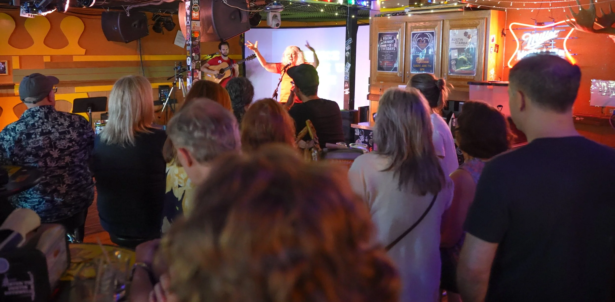 A lively live music performance at a bar with an audience enjoying the show. Zelda singing on stage, Dave Freeman beside her, and colorful stage lighting.