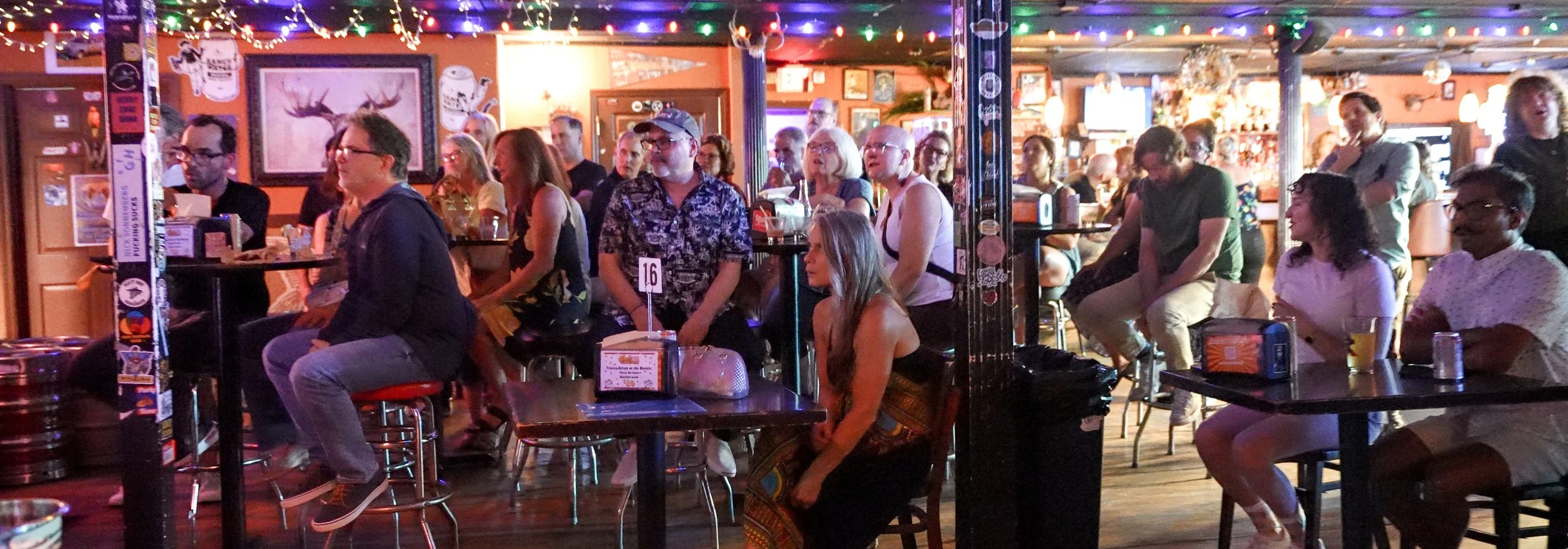 People sitting and standing at our Dive Bar Choir Launch Party, framed pictures, and stickers on the poles, with drinks on tables.