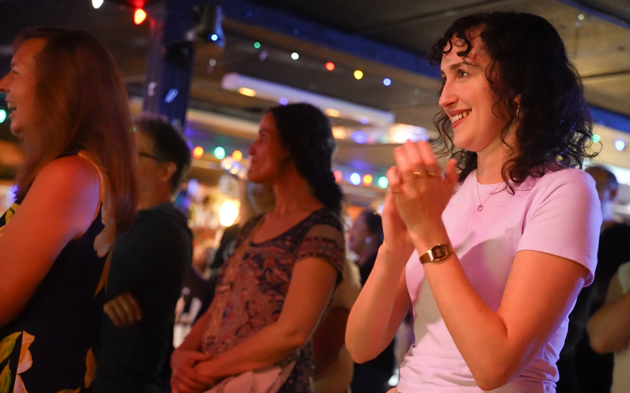 A group of women at our Dive Bar Choir Launch Party, standing close together, smiling, and clapping, with colorful string lights overhead.
