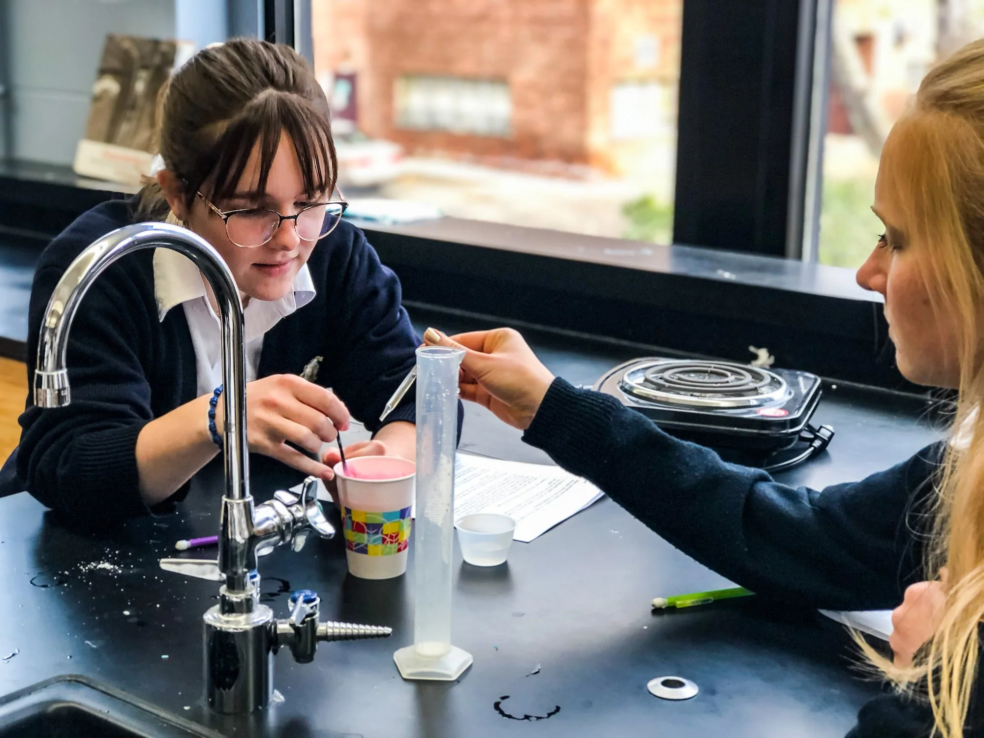 Two Chesterton Academy girls sitting at a science lab table with dishes, a test tube, and scientific equipment, working together in a classroom or lab setting.