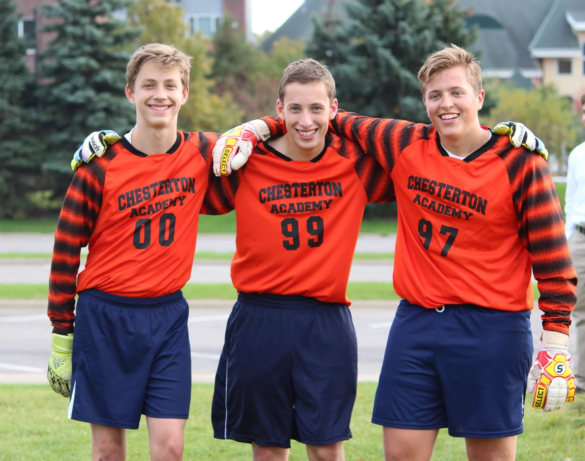 Three Chesterton Academy boys in orange soccer uniforms, standing on a grassy field.