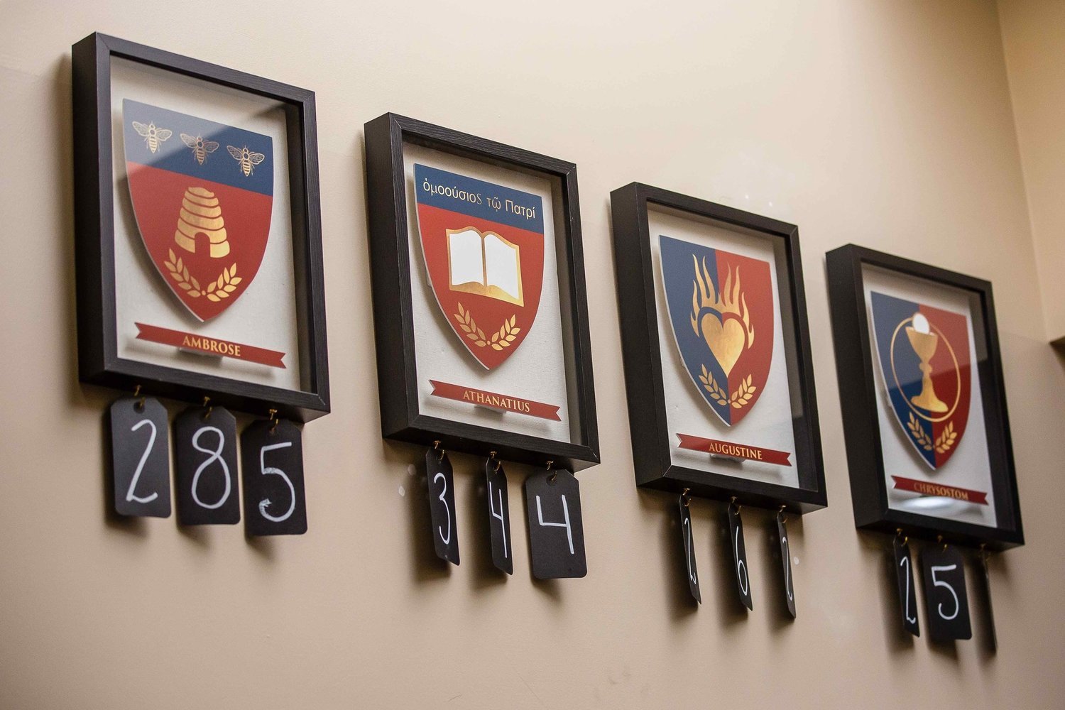 Four framed Chesterton Academy emblems on a wall, each with a banner and a black number tag hanging below, displaying 285, 34, 406, and 215. The emblems feature shields with different symbols and names.