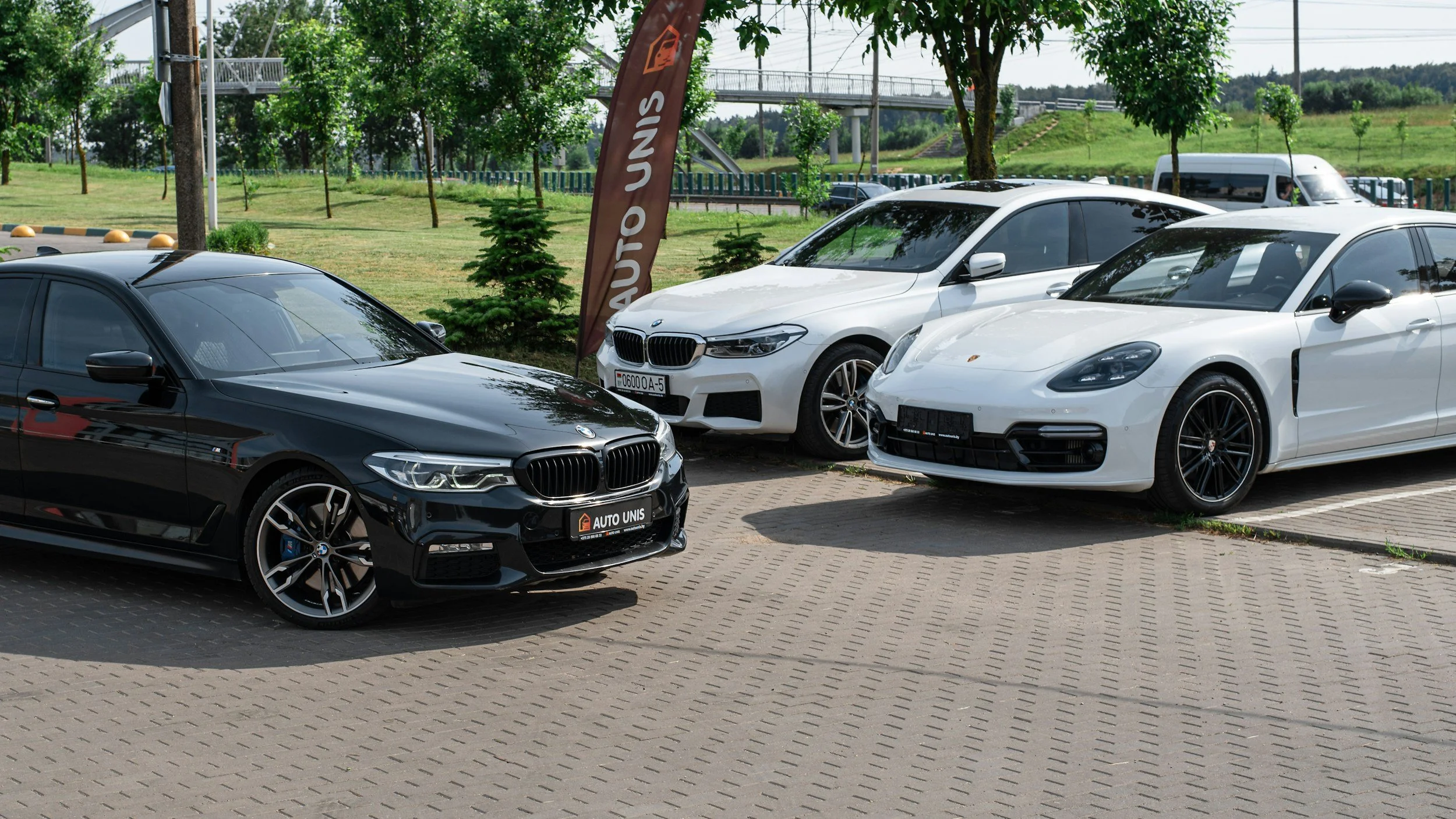 A parking lot with three white and black luxury cars, a black BMW, a white BMW, and a white Porsche, parked side by side. There is a brown banner with white text and a logo, and a green park with trees and a bridge in the background.