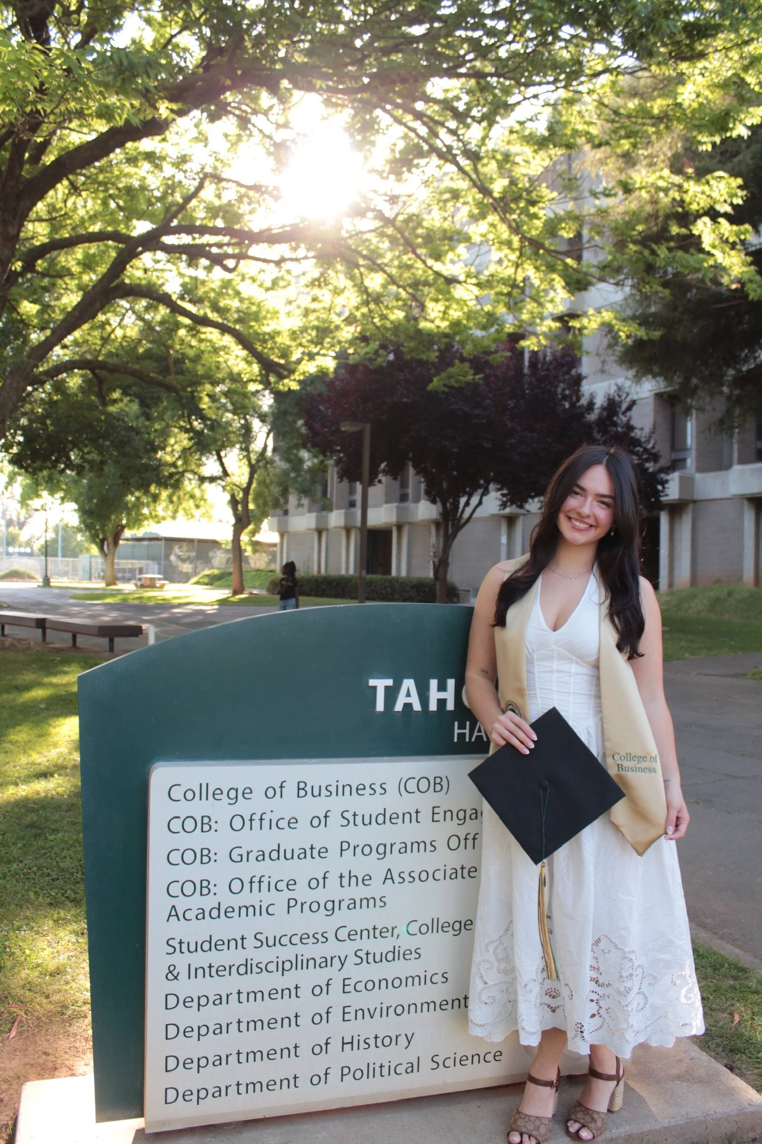 A young woman in a white dress holding a graduation cap standing next to a large sign for the College of Business at a university. She is smiling and outdoors on a sunny day with trees and a building in the background.