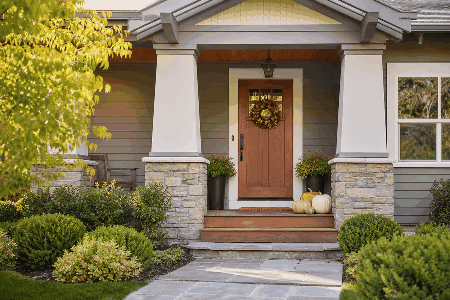 Front porch of a house with a wooden door decorated with a wreath, flanked by two potted plants, pumpkins on the steps, and surrounded by greenery and shrubs.