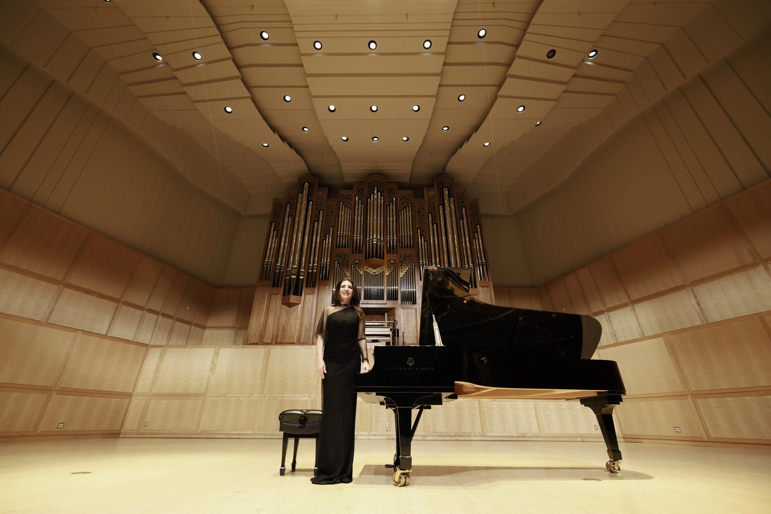 Female pianist standing next to a grand piano on a stage with wood-paneled walls and overhead lighting.
