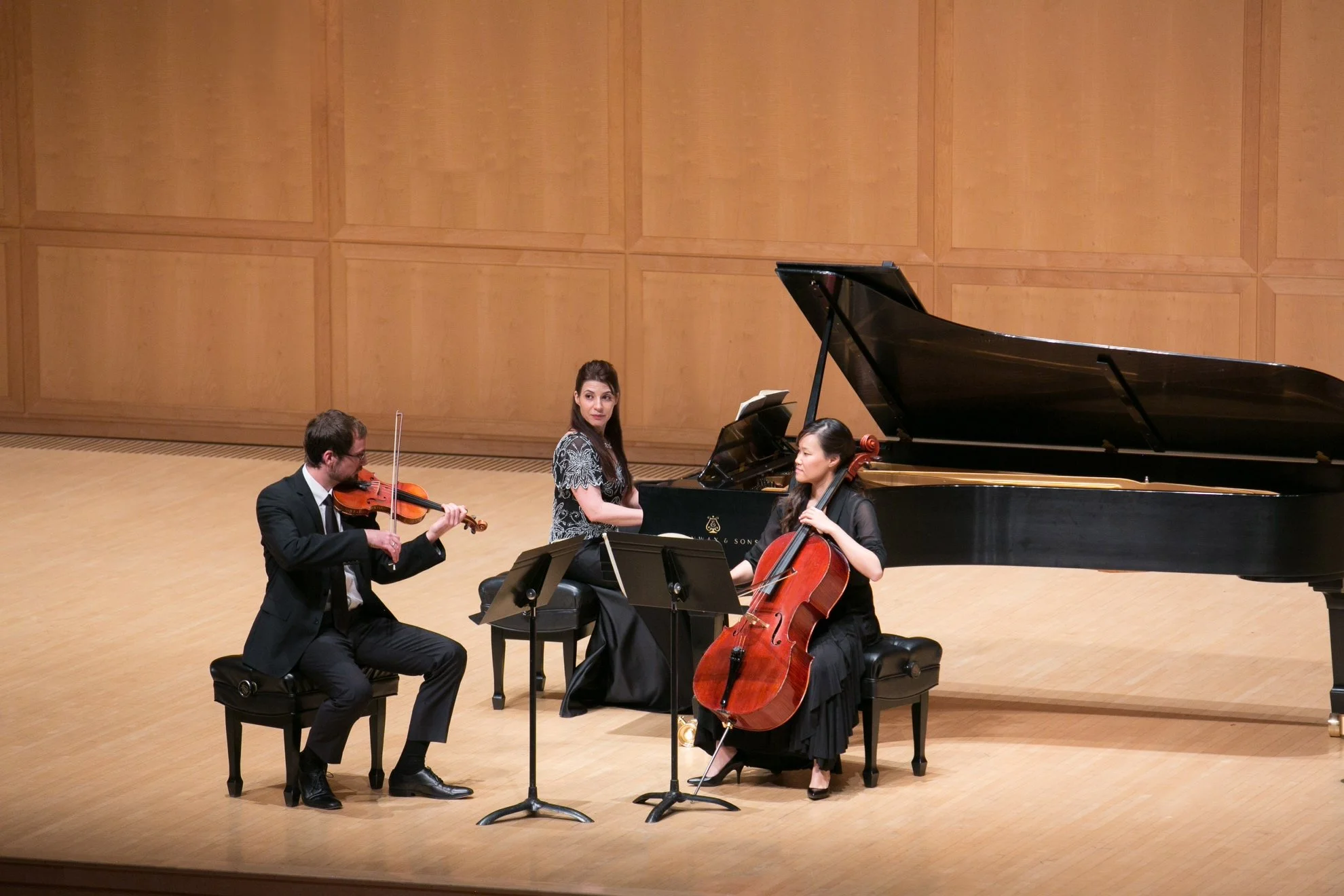 Four musicians performing on stage: a violinist, a pianist, a cellist, and a seated pianist. The stage has a wooden backdrop and a grand piano.
