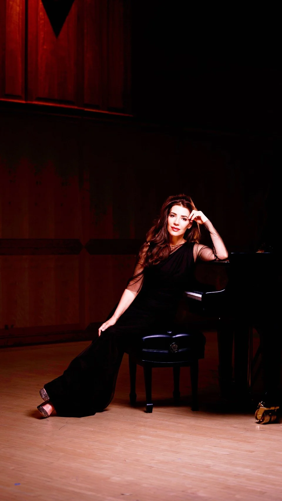 A woman sitting on a piano bench next to a grand piano, wearing a black gown with sheer sleeves, in a dimly lit room with wooden walls and floor.
