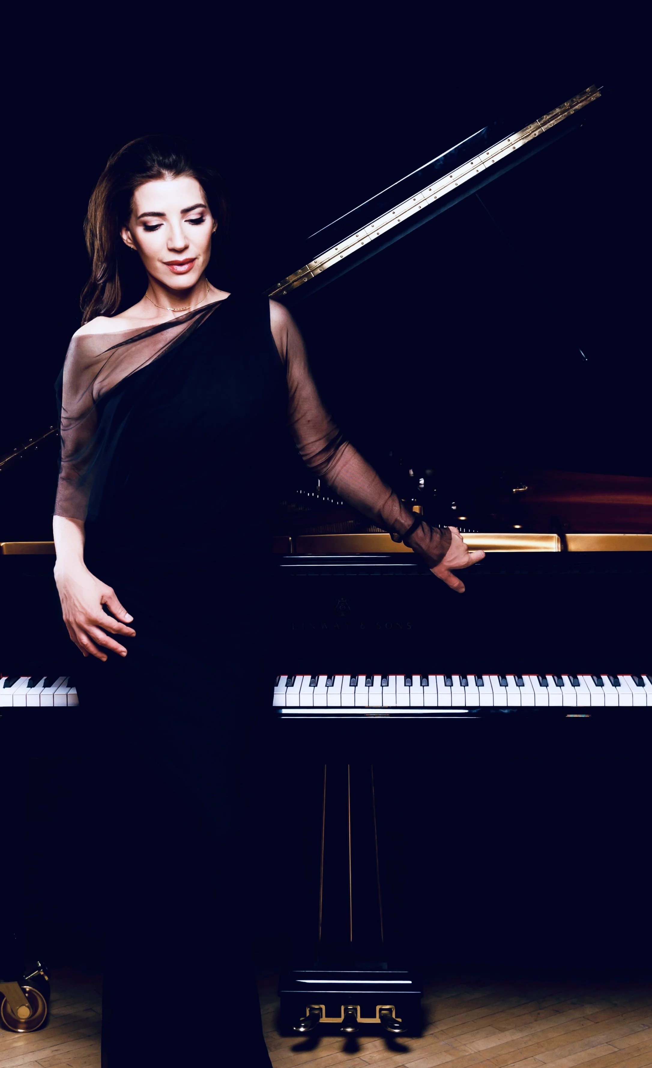 A woman in a black dress stands next to a grand piano with an open lid, looking down with closed eyes, in a dark setting with a wooden floor.