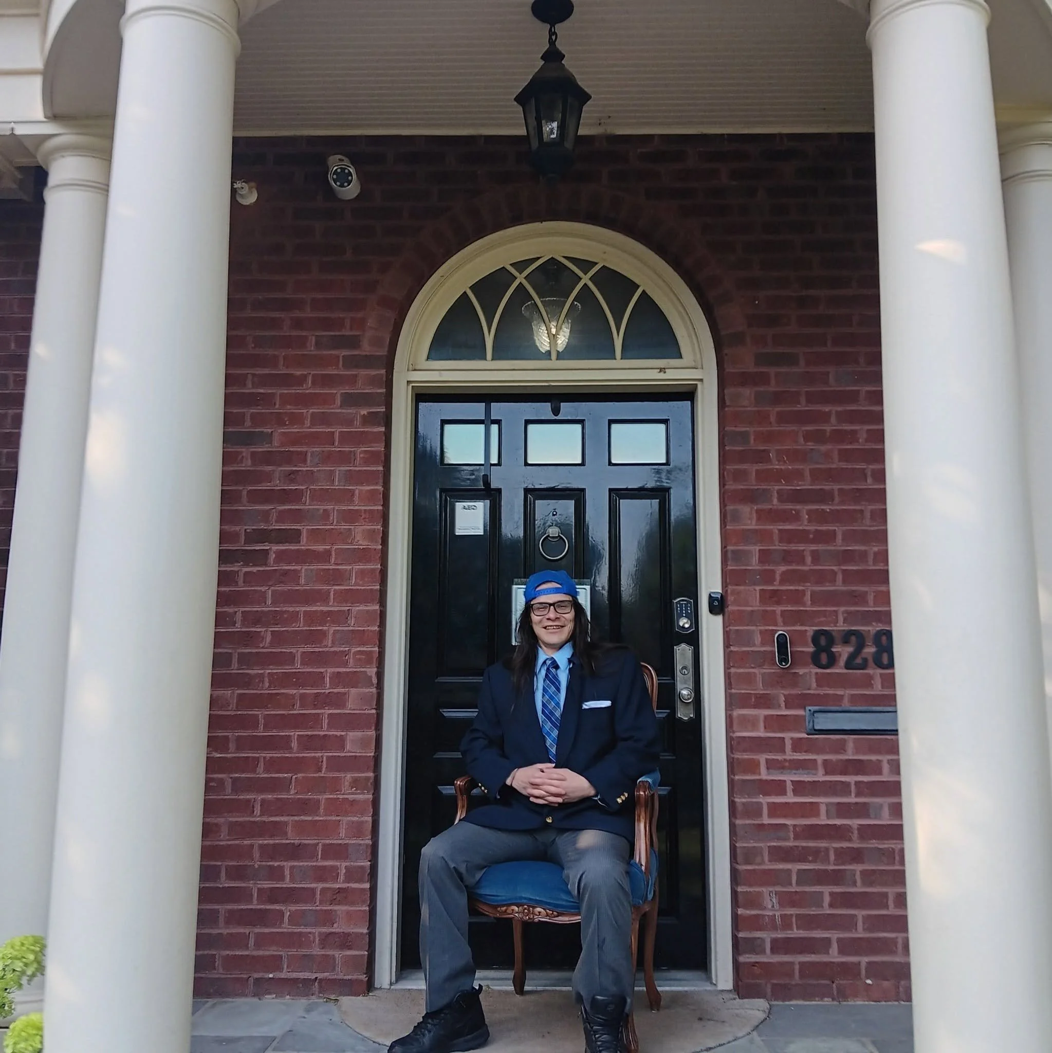A person in formal attire sitting in a chair in front of a brick house with white columns. The person is smiling, wearing a blue cap, glasses, a dark blazer, a white shirt, a blue tie, and dark pants.