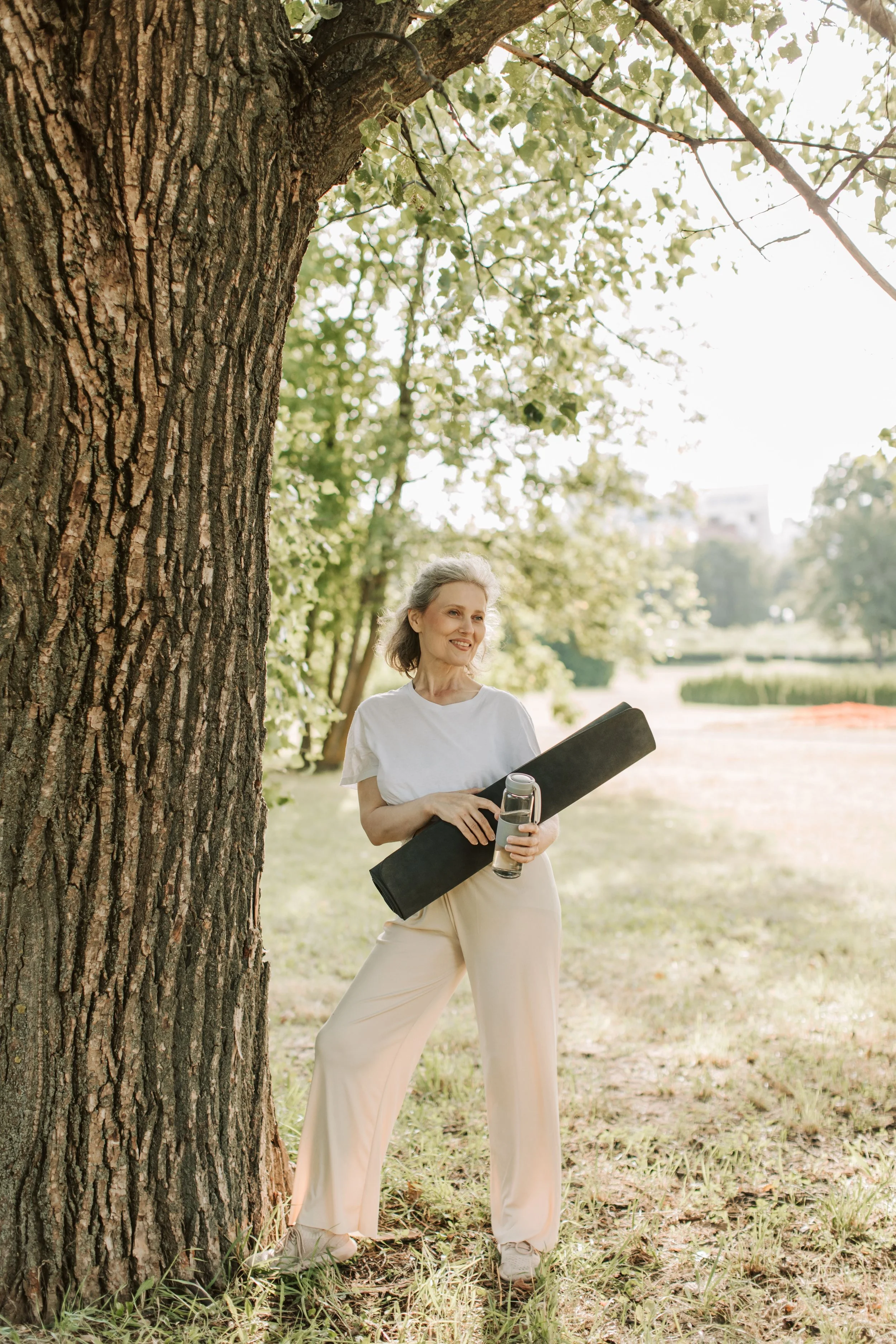 A woman standing outdoors in a park next to a large tree, holding a water bottle and a yoga mat, with a smile on her face, wearing casual light-colored clothing, under sunlit green foliage.
