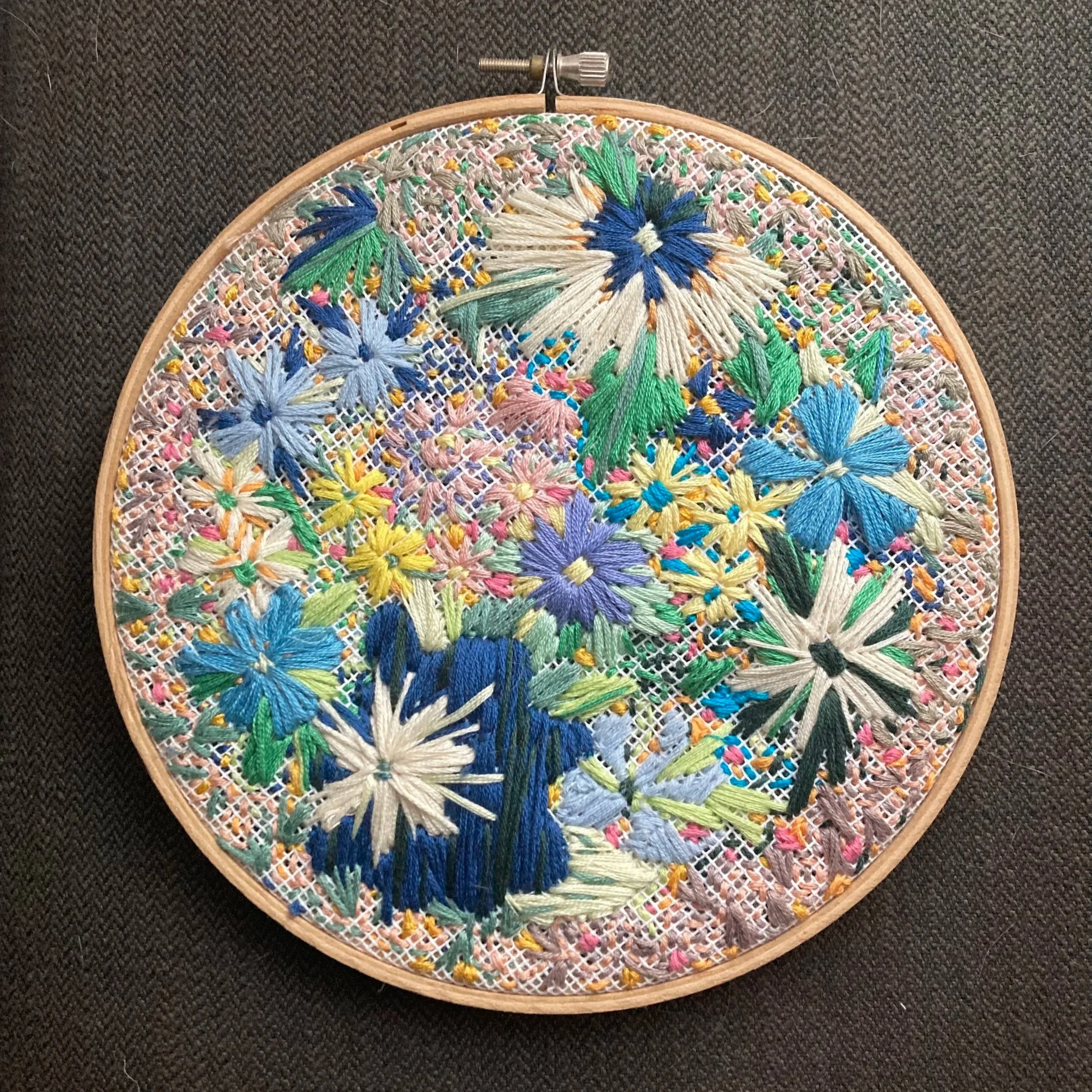 A colorful embroidery with floral patterns in various shades, housed in a round wooden hoop on a dark textured surface.