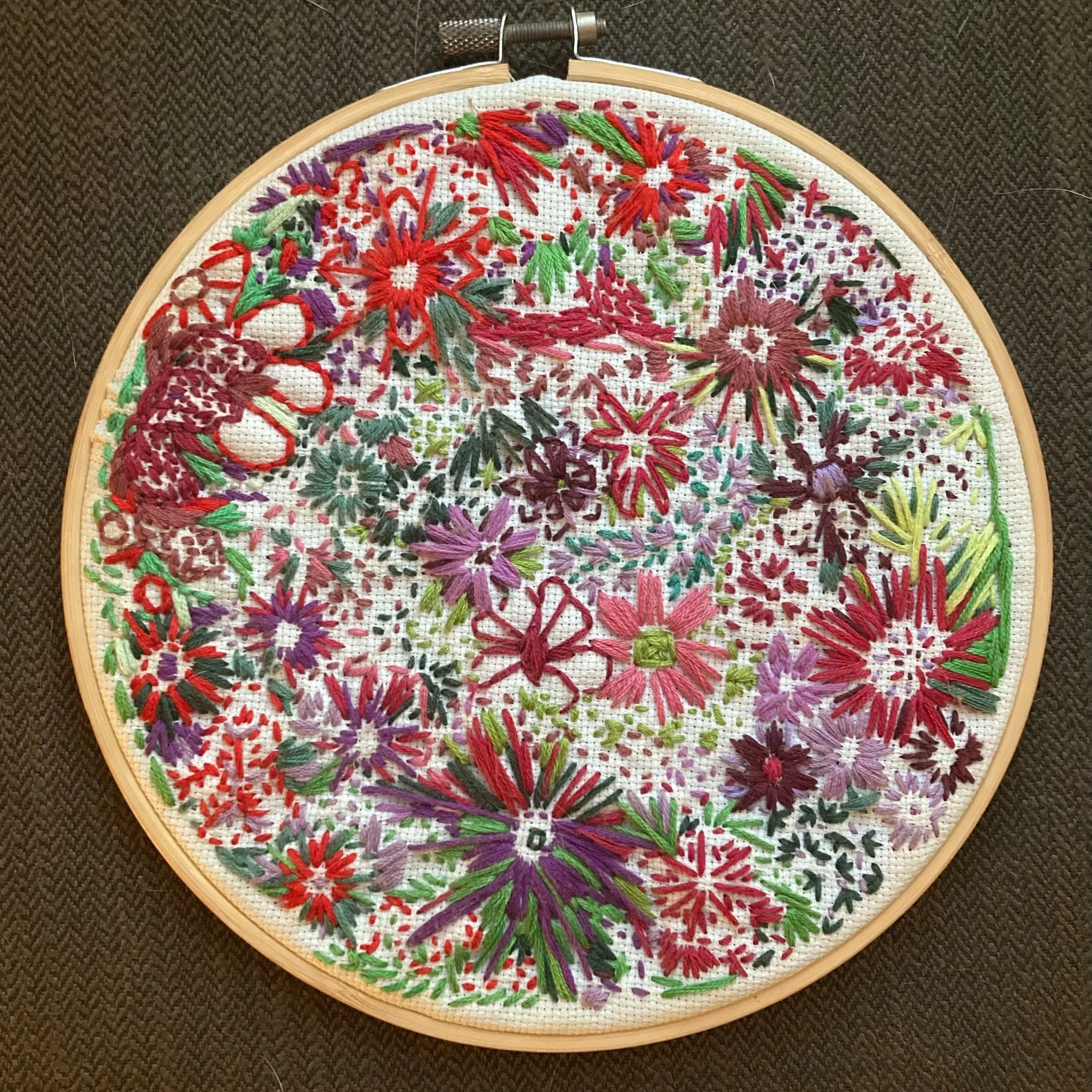 A colorful embroidery of a bouquet of flowers with red, purple, yellow, and green threads on a white fabric, stretched in a wooden embroidery hoop on a dark textured surface.