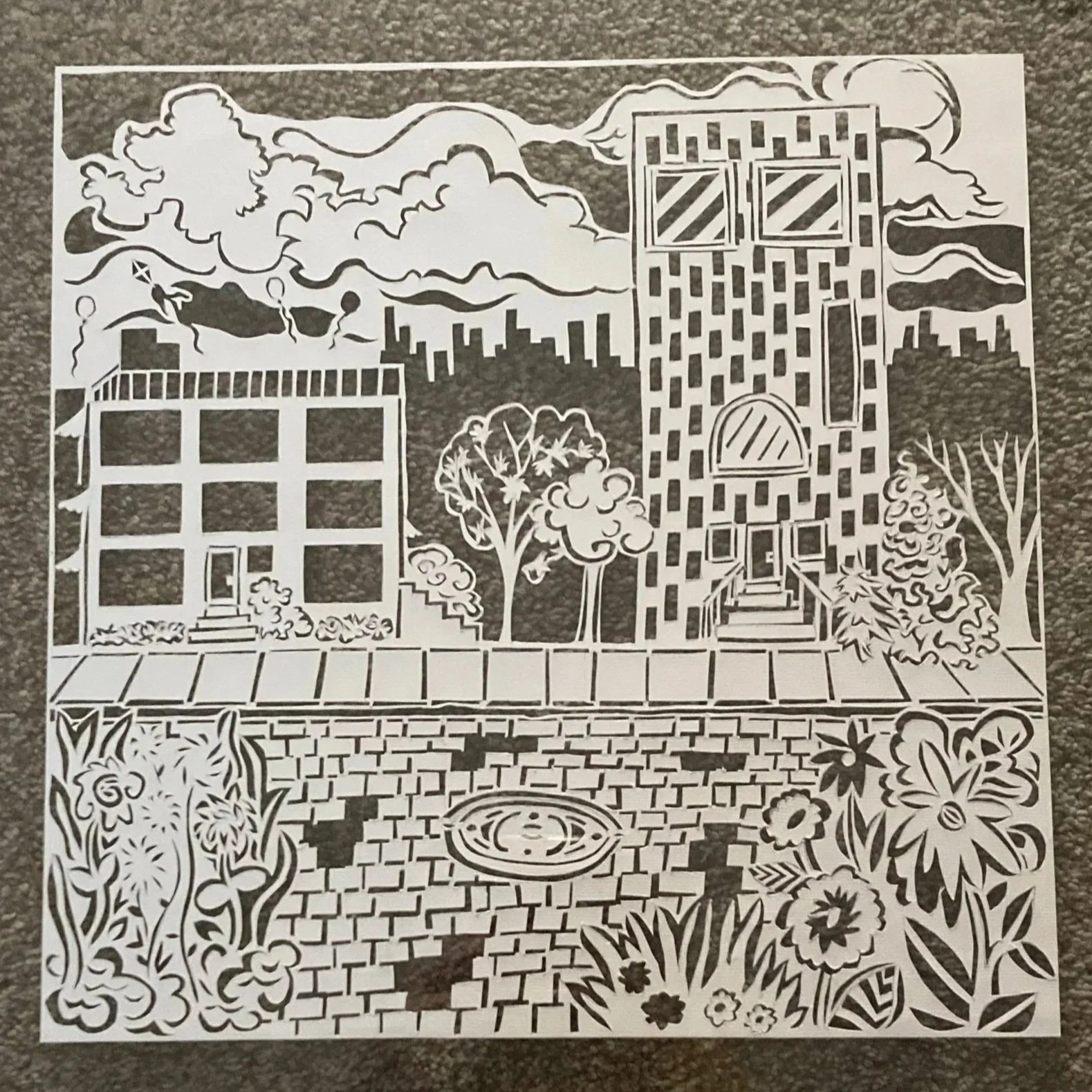 Black and white paper cutout of a city scene with buildings, trees, a fountain, and decorative plants, laid on a textured gray surface.