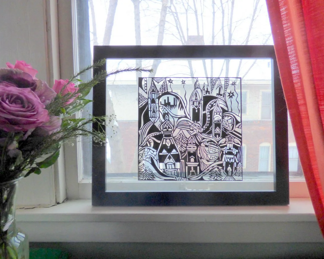 Decorative paper cutout art of a cityscape with castles, mountains, and stars, placed on a windowsill with a vase of pink and purple flowers to the left, and red curtains on the right.