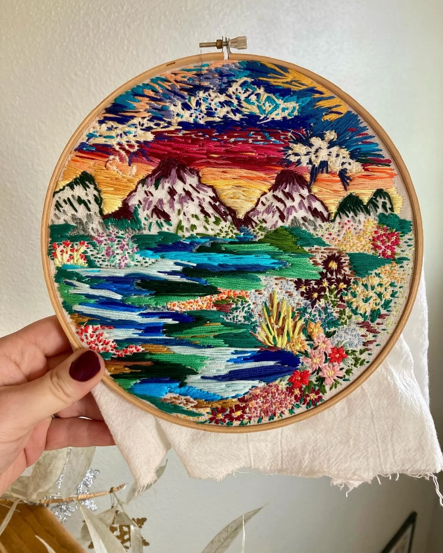 Embroidery hoop with a landscape scene featuring mountains, a sunset, a river, and various colorful flowers.