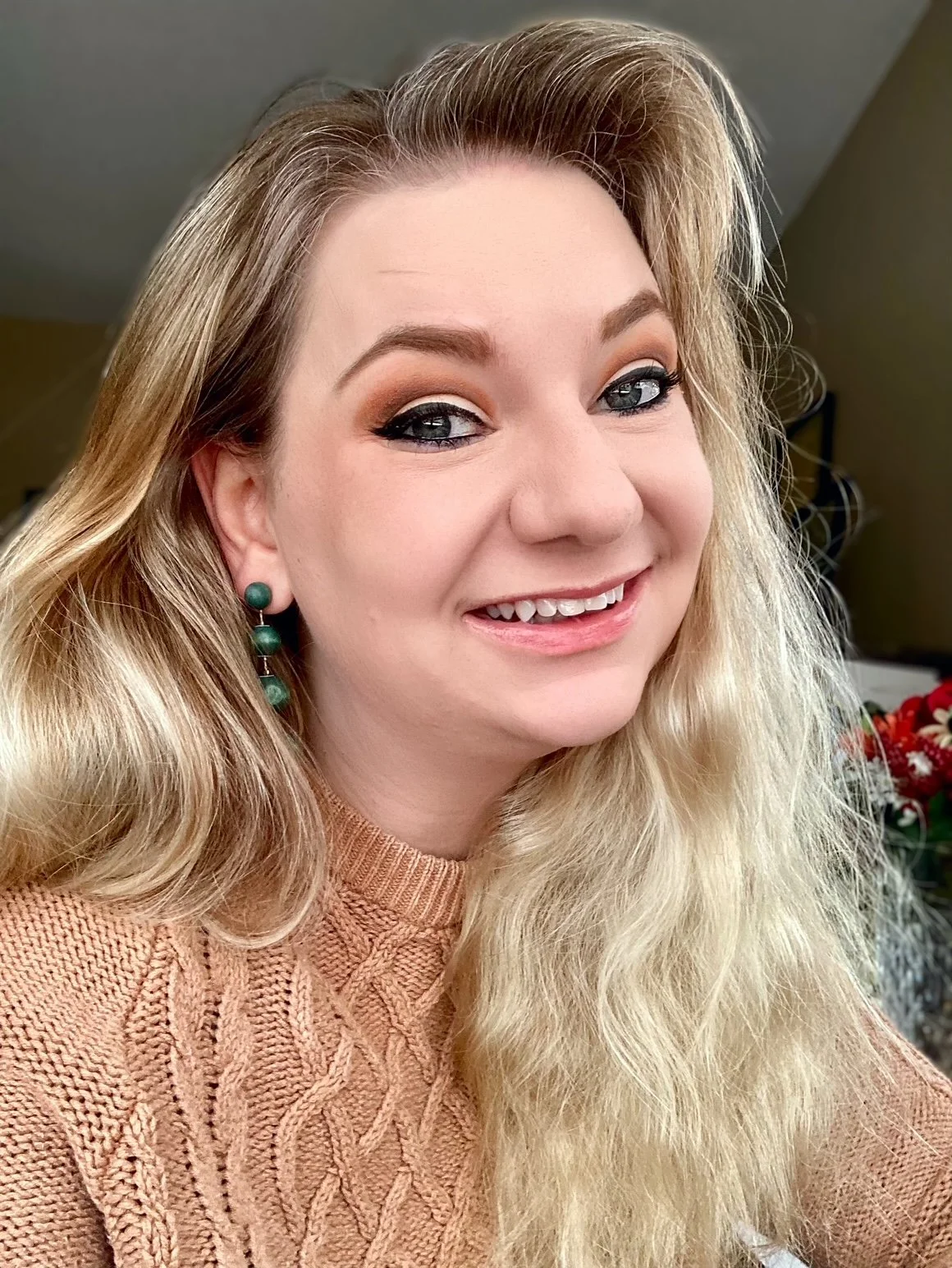 A woman with long, wavy blonde hair, wearing a peach-colored sweater and green earrings, smiling indoors.