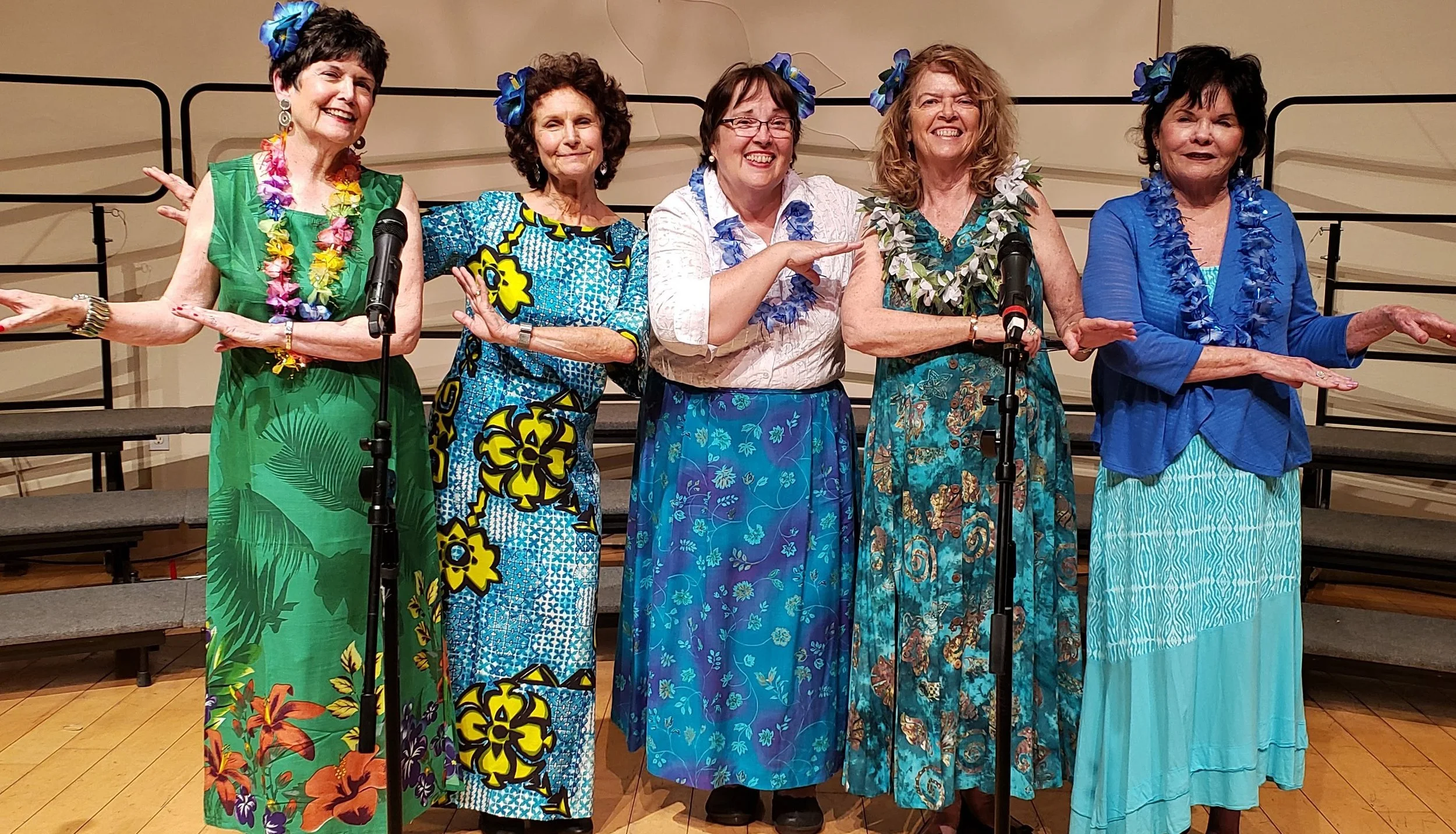 Five women standing on a stage, dressed in colorful Hawaiian-style dresses and leis, performing in what appears to be a musical or choir group. They are smiling and gesturing with their arms.