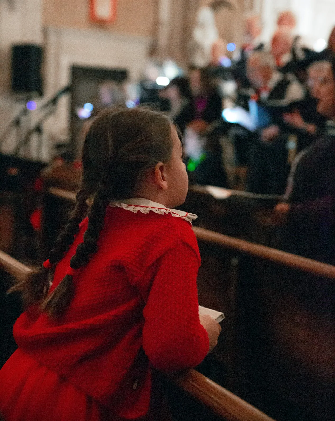 A young girl with braided hair wearing a red dress sitting in a church or concert hall during a performance or event, with an audience in the background.