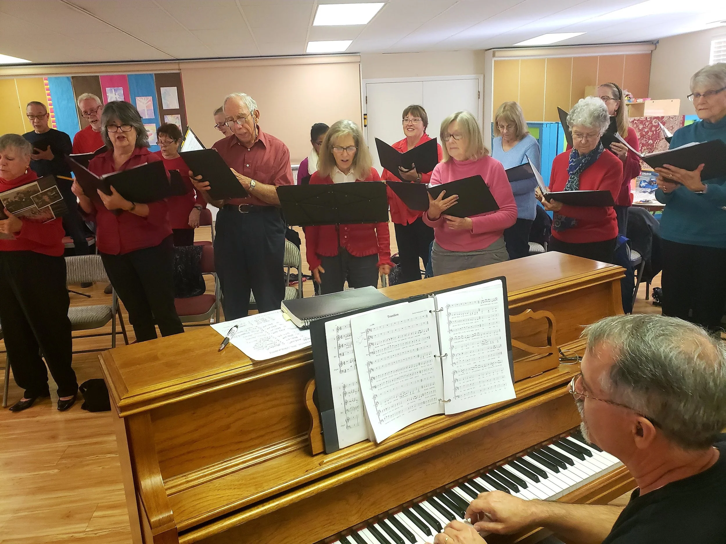 A group of elderly people singing in a choir, standing behind a wooden piano with sheet music, in a room with colorful walls and decorations.