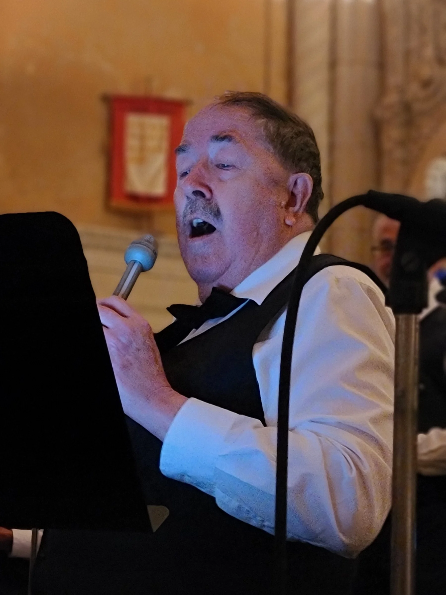 A middle-aged man with short, dark hair and light skin is speaking into a microphone at a formal event. He is wearing a white shirt and a black vest, and appears to be in a room with ornate wood paneling and warm lighting.