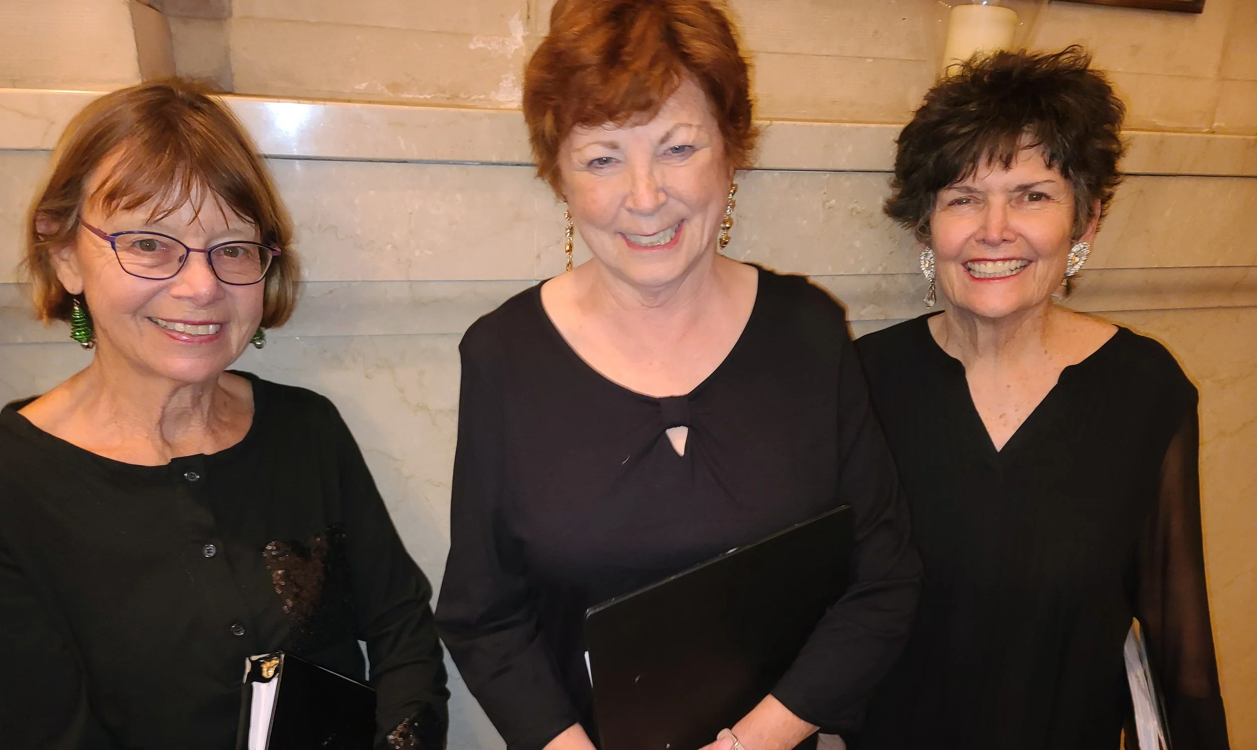 Three women dressed in black, standing in front of a beige stone wall. The women are smiling, with the woman in the middle having red hair and wearing drop earrings, while the women on each side have glasses and earrings.