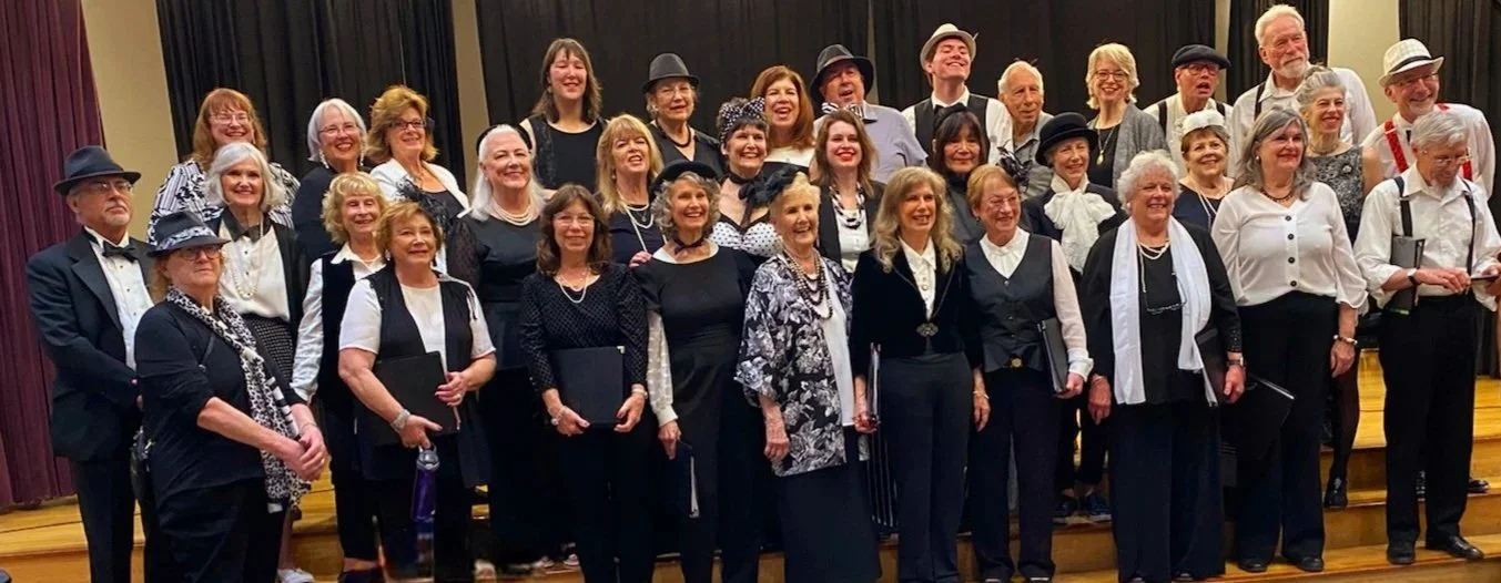 Group of smiling adults posing on a stage, dressed in vintage or retro attire, possibly for a performance or celebration.