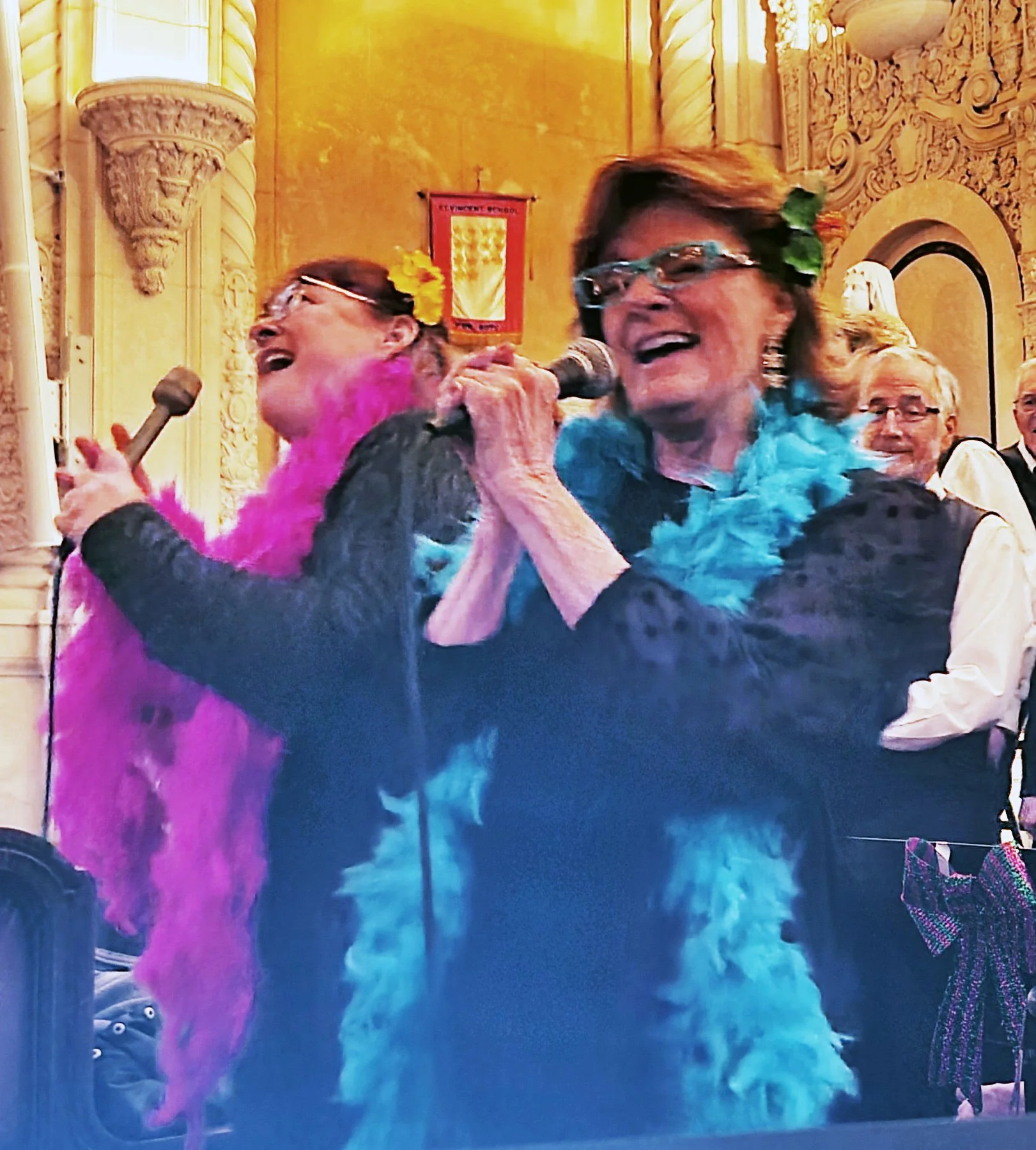 A group of women at a celebration, wearing colorful feather boas and floral accessories, singing into a microphone in an ornate room with gold-colored walls and decorated columns.