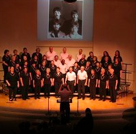 Novato Music Association Chorus performing on stage with a conductor in front. The chorus members are dressed in black, and there is a screen above showing a black-and-white photo of three children.