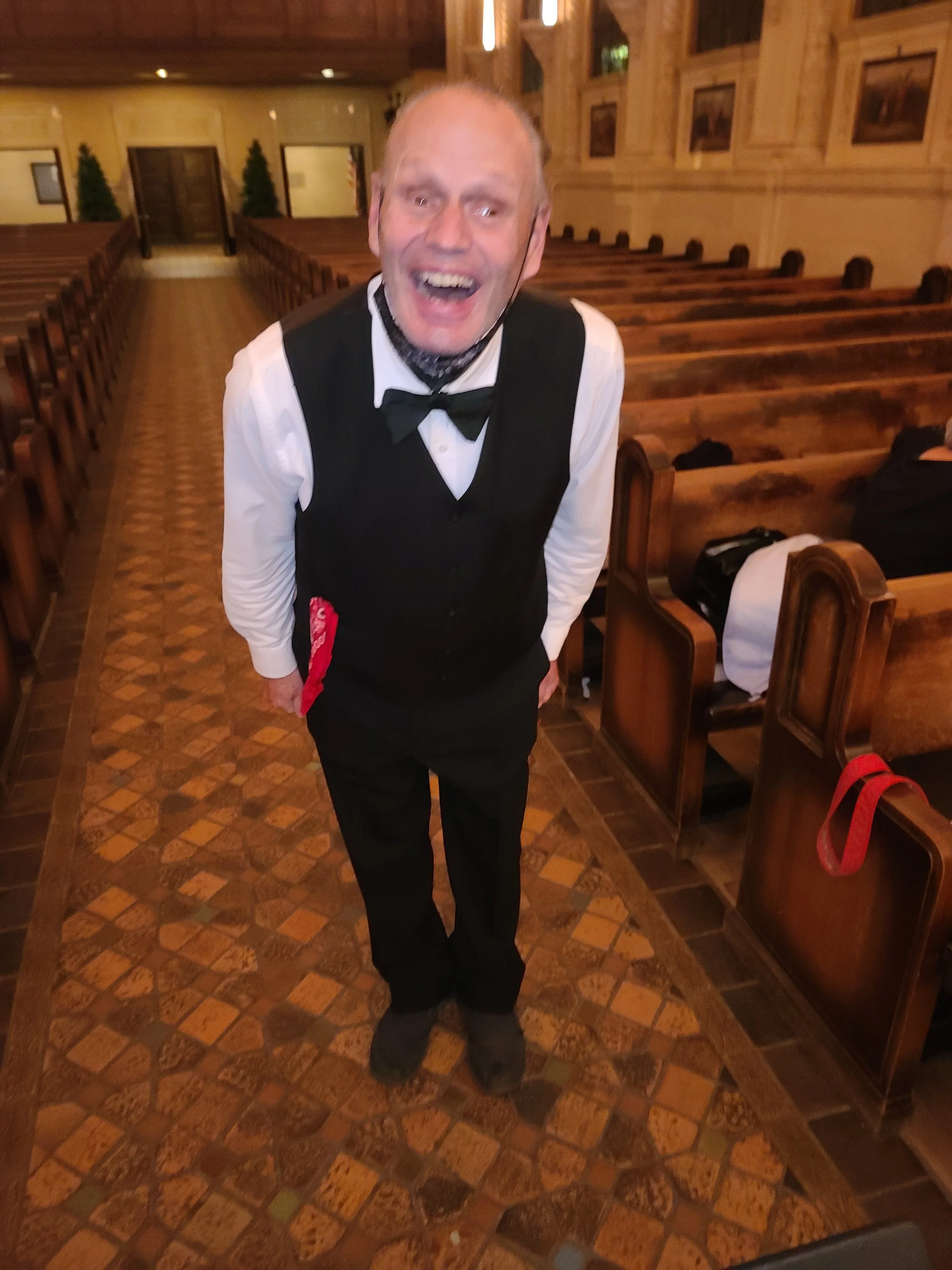A man dressed in formal attire, including a vest and bow tie, stands inside a wooden church or chapel, smiling or laughing, with pews and wooden paneling visible in the background.