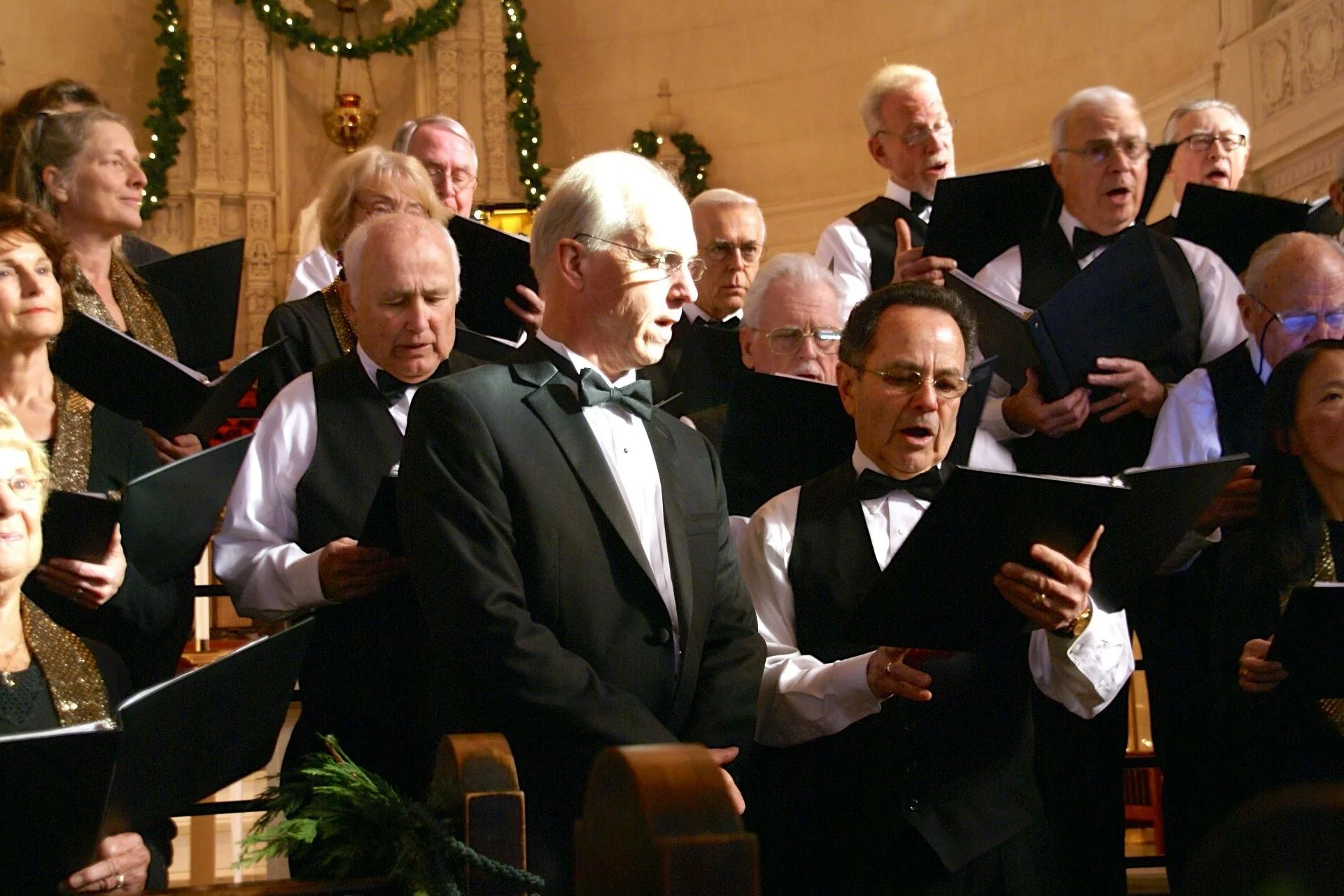 A group of senior choir singers in formal black and white attire singing in a church, decorated with Christmas garlands and ornaments.