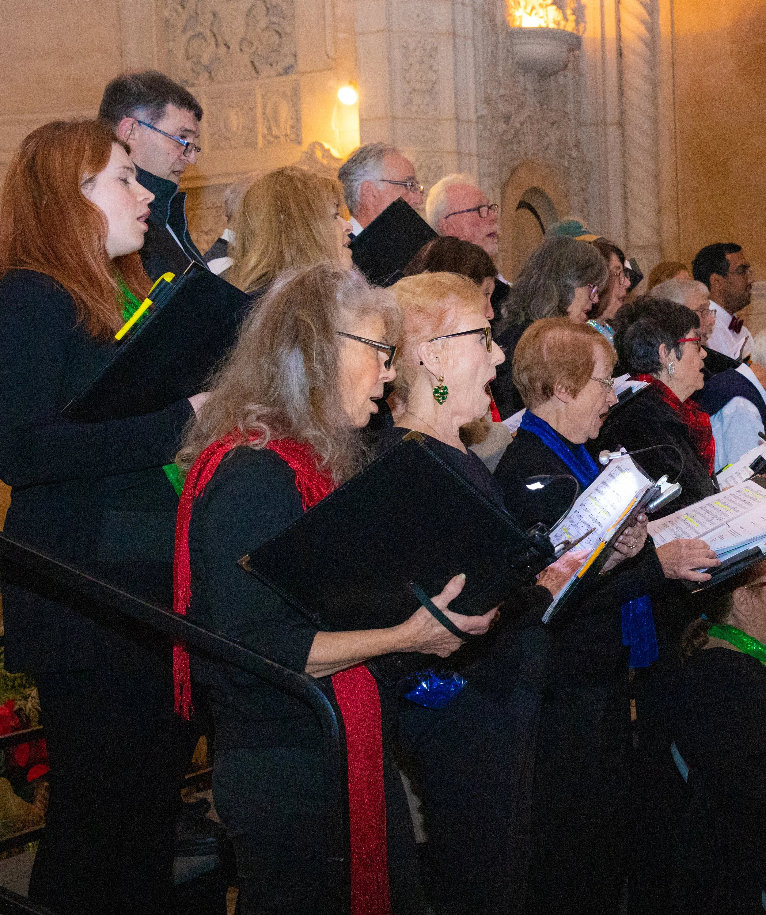 Group of people singing or performing in a concert hall, holding music sheets or songbooks, with ornate architectural details and warm lighting in the background.
