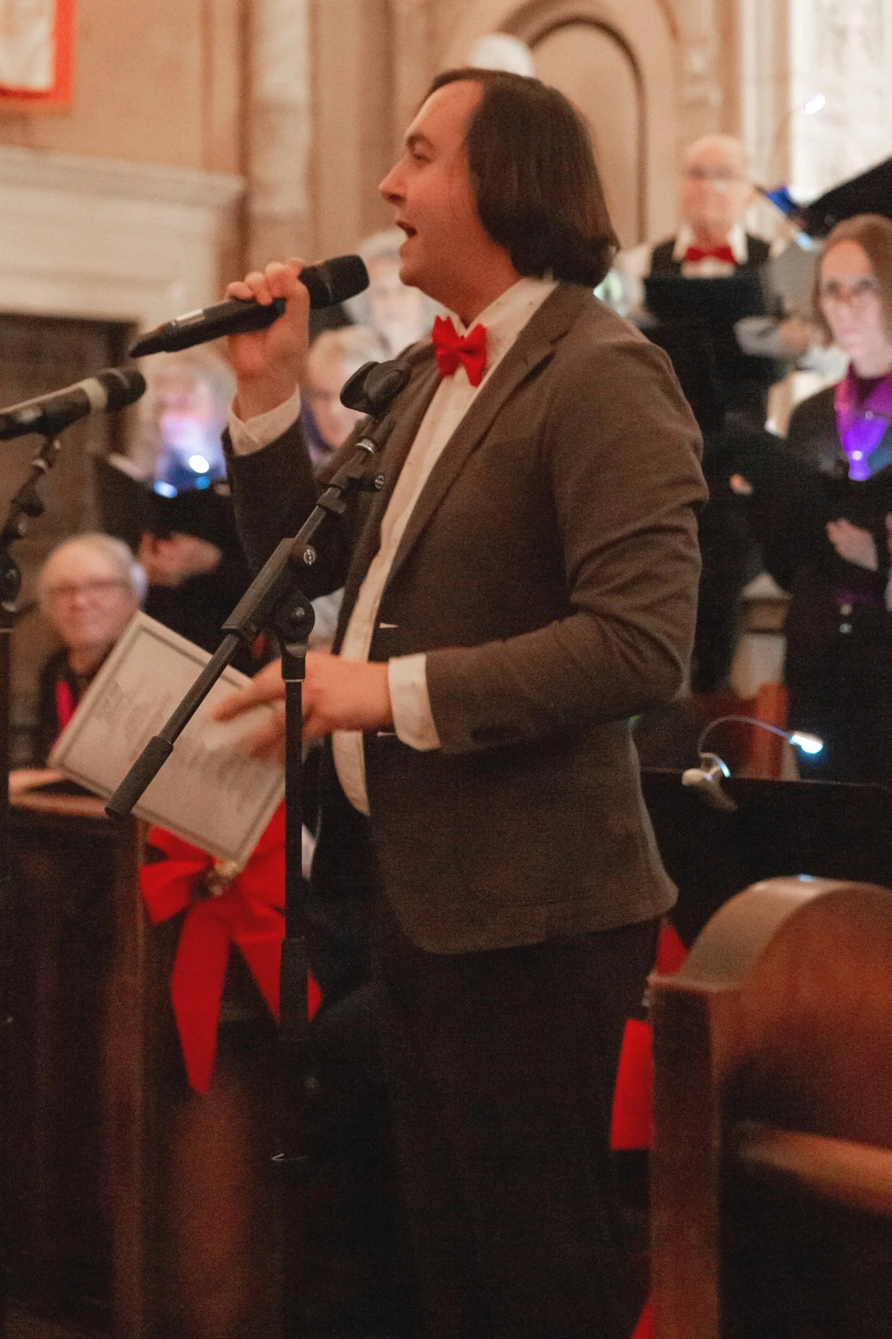 A man with dark hair wearing a gray suit and red bow tie singing into a microphone during a performance in a formal setting with an audience.