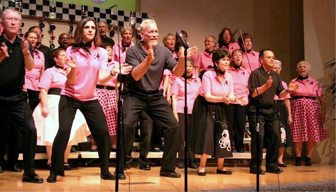 A group of men and women on stage, singing and dancing with microphones, wearing coordinated pink and black outfits, in a room decorated with Christmas ornaments.