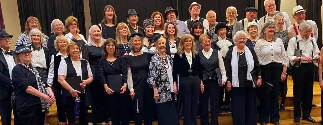 Group of elderly men and women dressed in formal and semi-formal attire on stage, likely after a choir performance, smiling and posing for a photo.